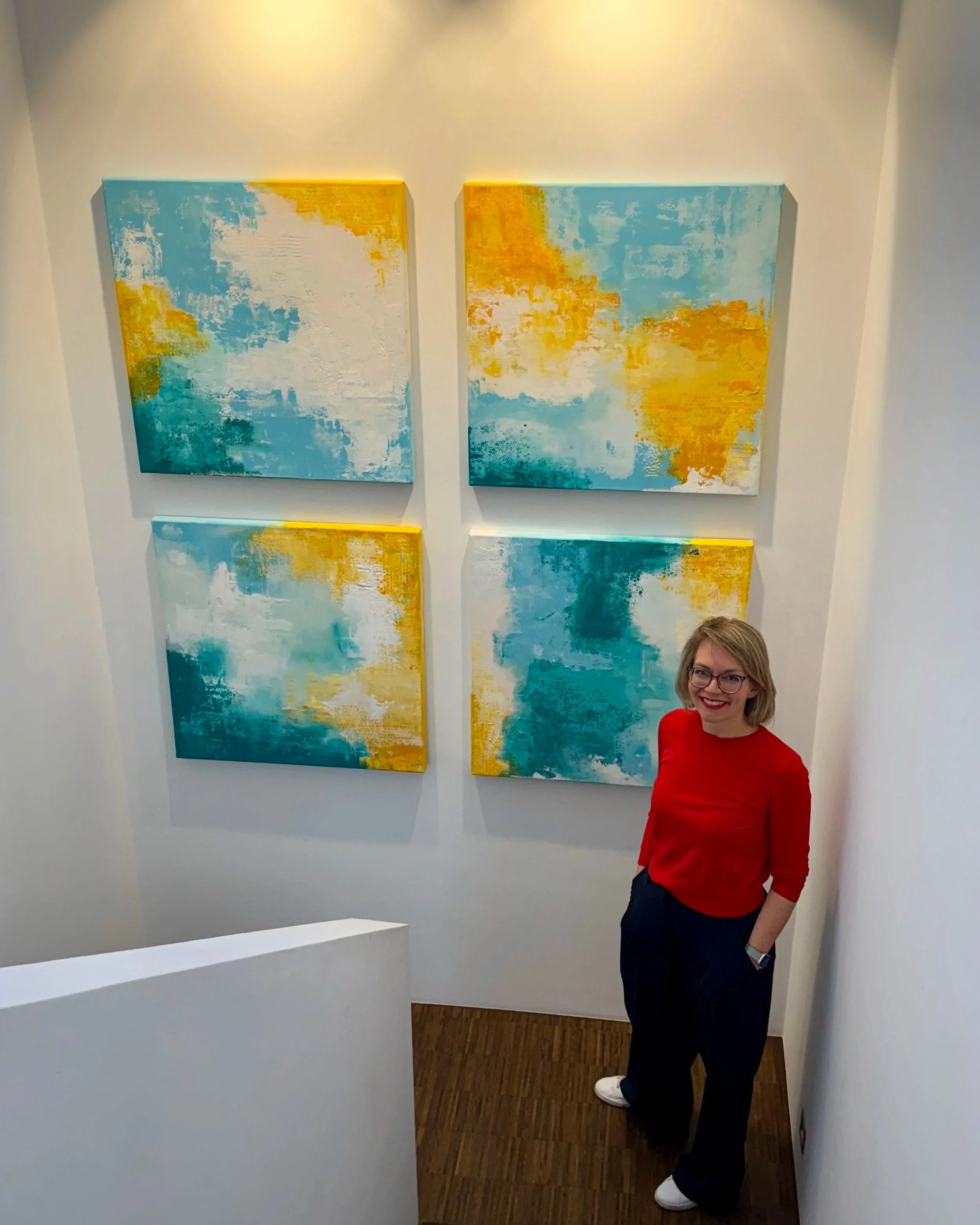 Eine Frau mit Brille und braunen Haaren, die in einem roten Oberteil gekleidet ist, steht in einem Raum vor einer weißen Wand. Über ihr hängen vier abstrakte Gemälde in Blau, Weiß und Gelb. Die Frau lächelt und schaut in die Kamera.