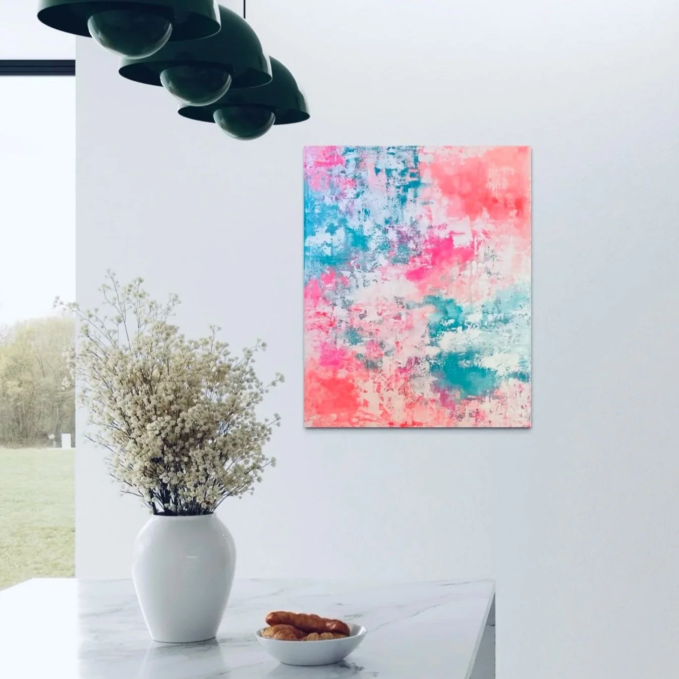 Moderner Raum mit Wandbild in Pink und Blau, weiße Vase mit weißen Blumen auf Marmor-Tisch, Schüssel mit Gebäck, schwarzer Lampenschirm, Blick nach draußen auf grünen Rasen und Bäume.