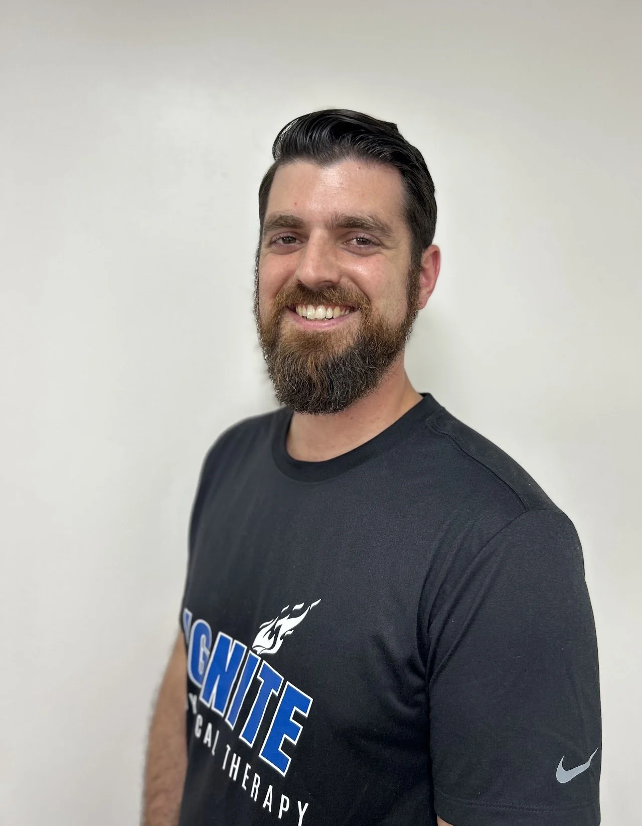 A smiling man with a beard and dark, styled hair, wearing a black sports T-shirt with a logo and text, standing against a plain light-colored wall.