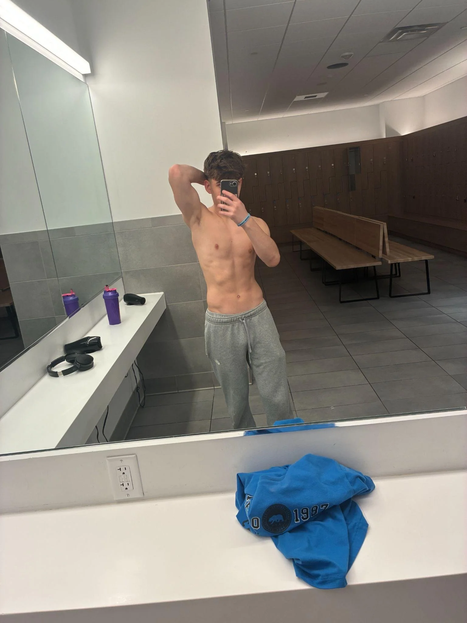A shirtless young man with brown hair standing in front of a mirror in a locker room, taking a selfie. The locker room has wooden lockers, benches, and gray tiled floors. There are purple bottles, headphones, and a blue shirt on the counter.