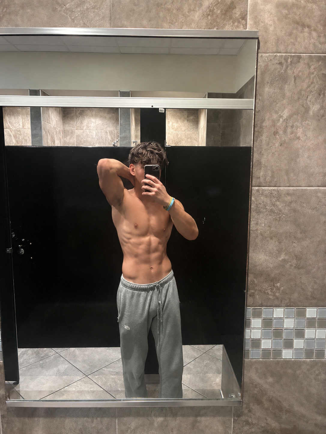 A young man taking a mirror selfie in a gym locker room, shirtless and wearing gray sweatpants, showing defined abs and arm muscles.