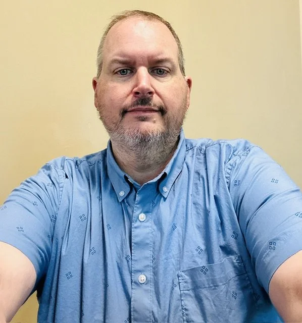 A man with a beard and short hair taking a selfie against a plain yellow background, wearing a blue button-up shirt.