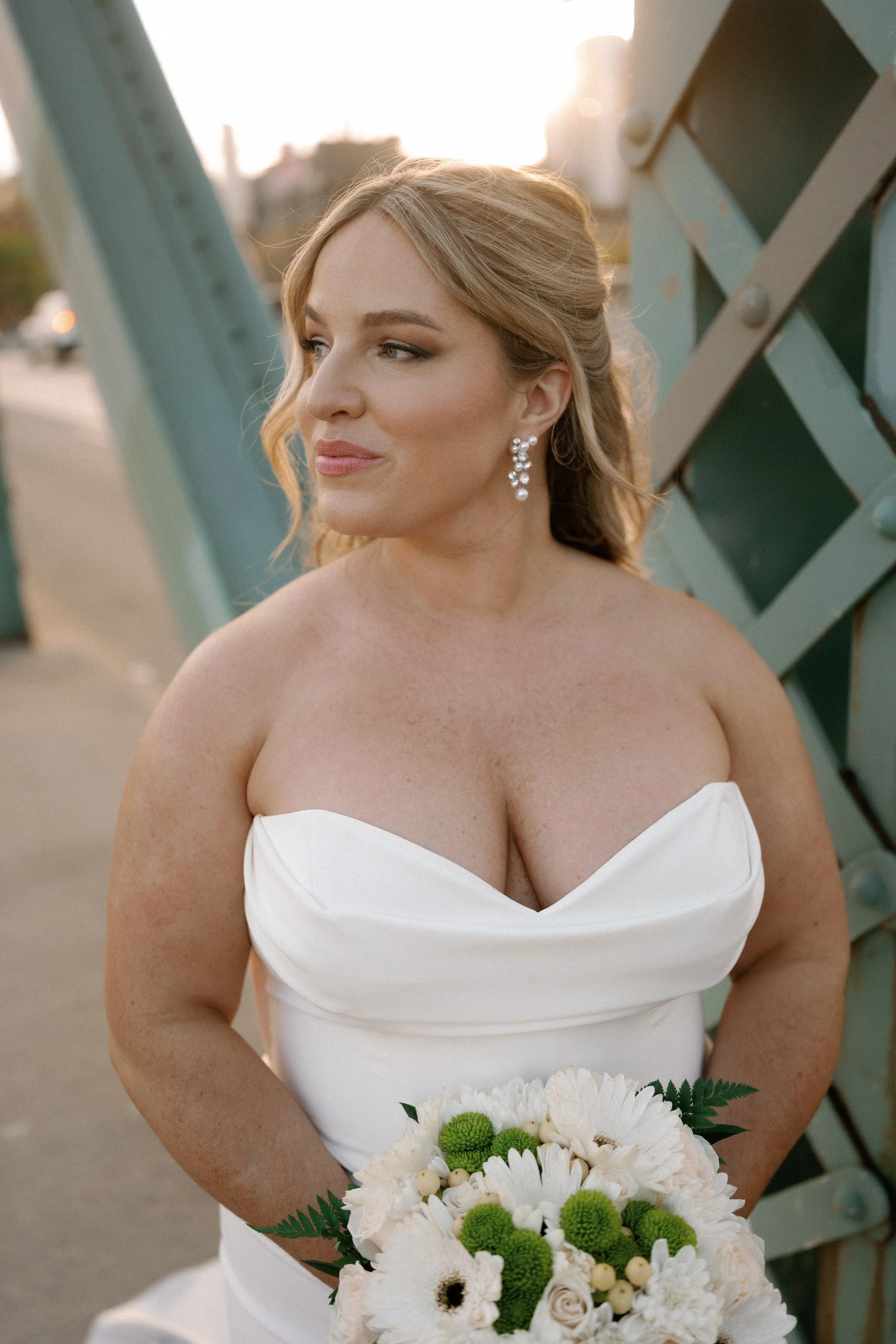 A woman in a white strapless wedding dress holding a bouquet of white and green flowers, outdoors during sunset.