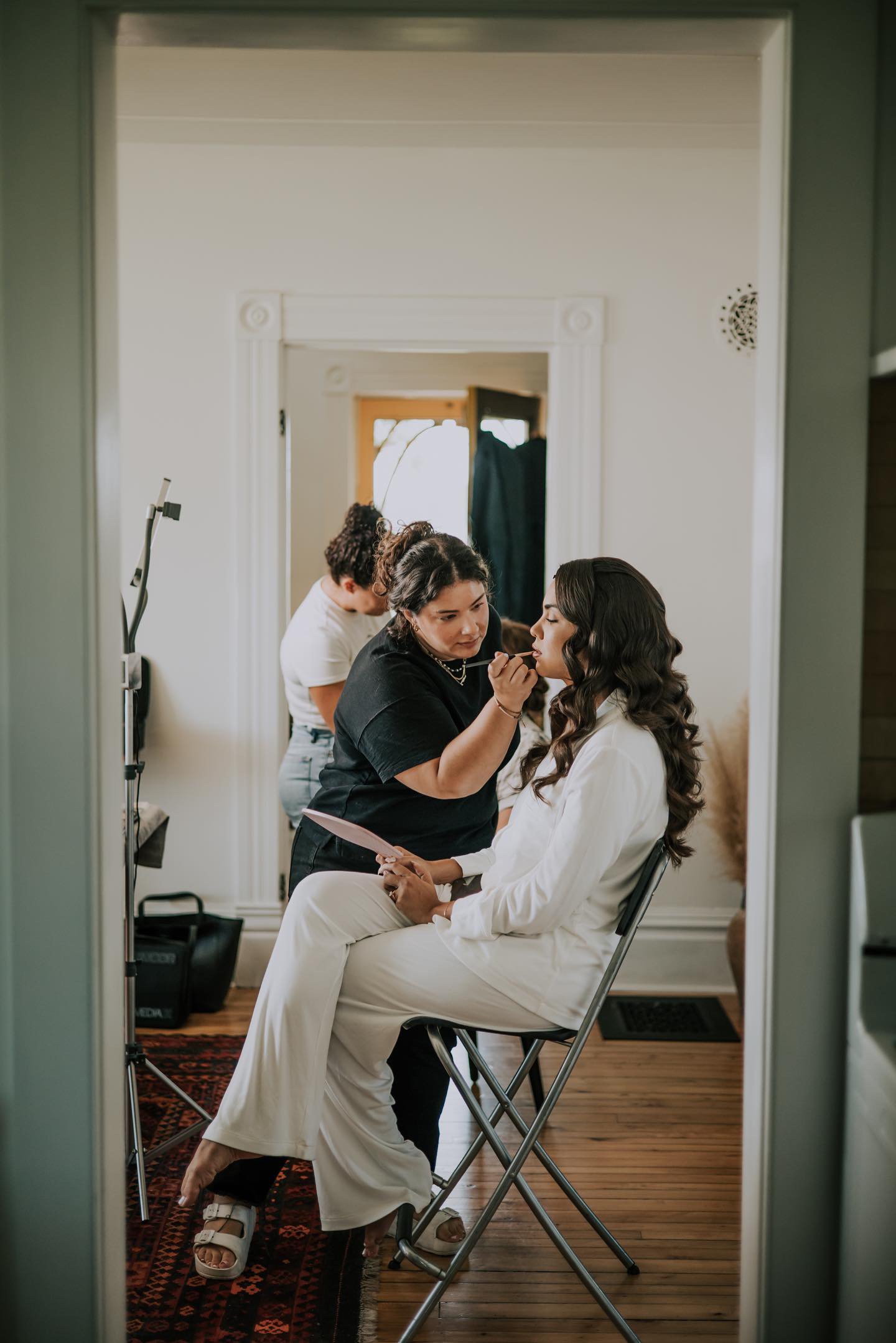 A woman is seated in a rehearsal room, having her makeup or hair done, with a makeup artist attending her. The woman is dressed in a white outfit and is holding a mirror or brochure. The room has hardwood floors and a patterned rug, with a doorway and a small piece of equipment visible in the background.