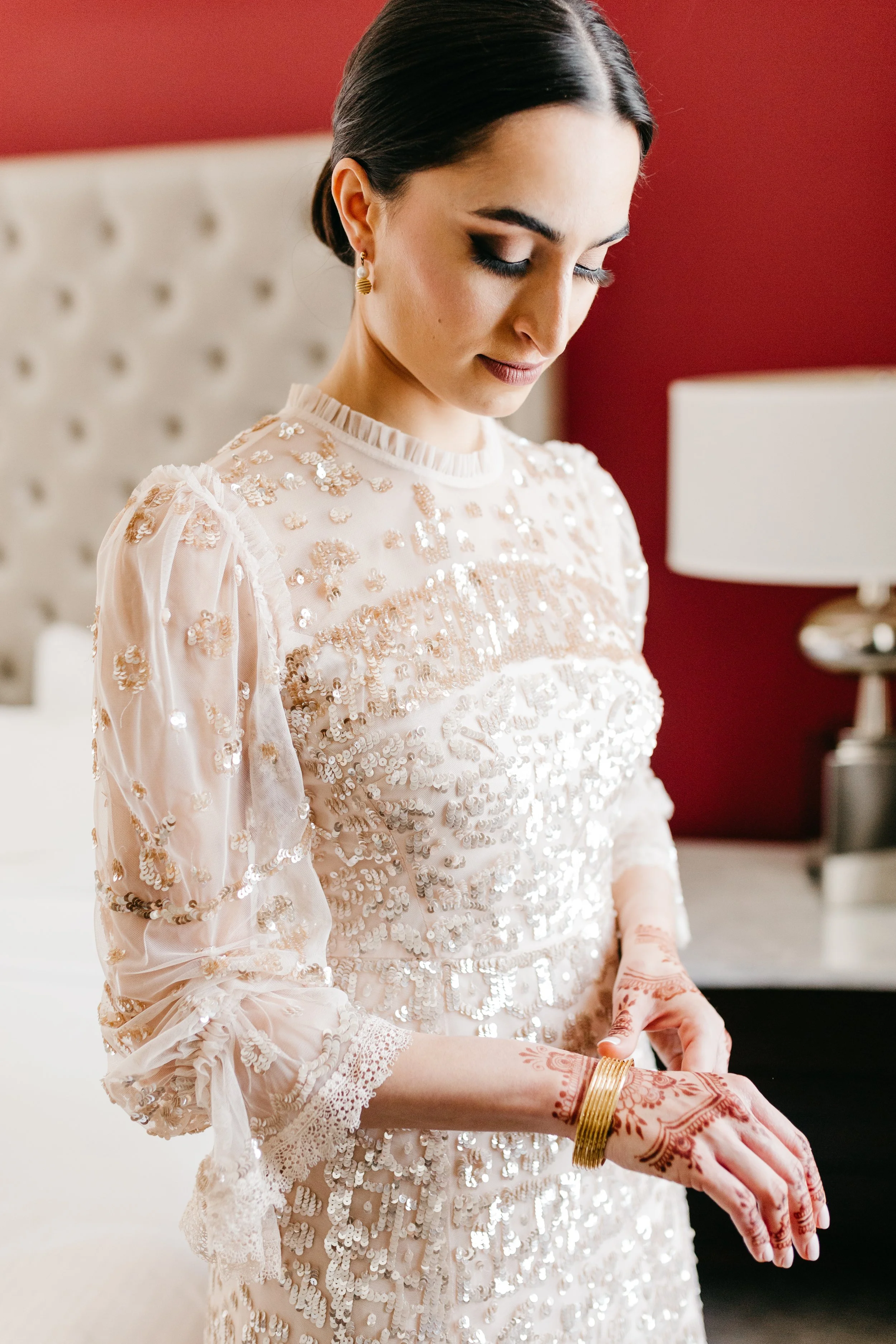 A woman in traditional Indian attire, wearing a cream-colored dress with sequins and embroidery, showing henna tattoos on her hands and gold bangles, with earrings, in a room with a red wall and a white bed in the background.
