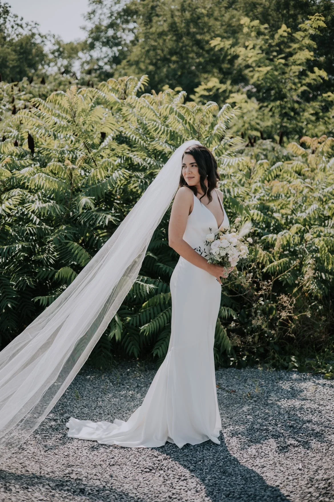 Bride with long white veil and natural makeup standing in front of trees with a white bouquet