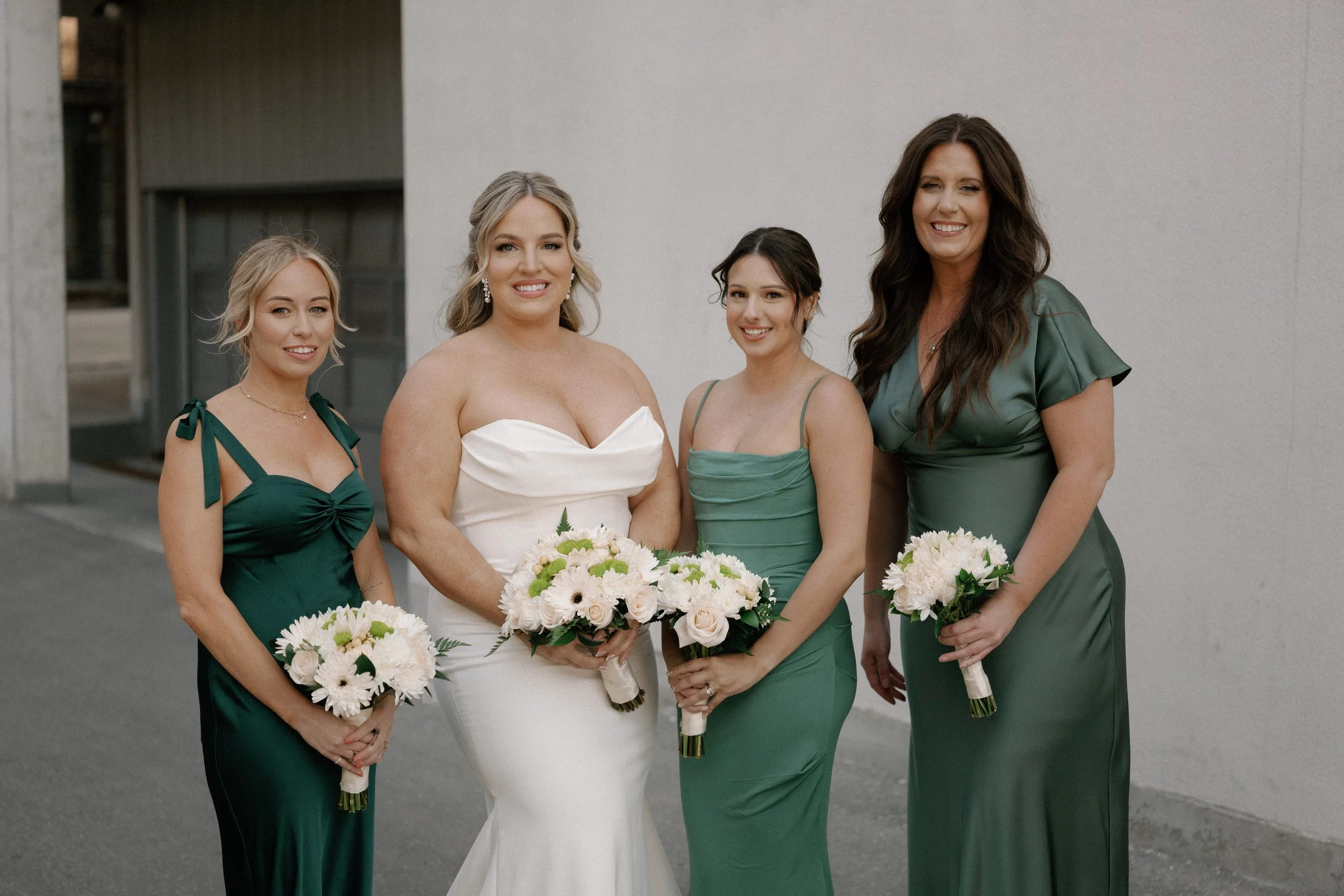Group of four women dressed in formal attire, holding bouquets of flowers, standing against a plain wall. One woman is wearing a white dress, indicating a wedding, while the others are in green dresses.