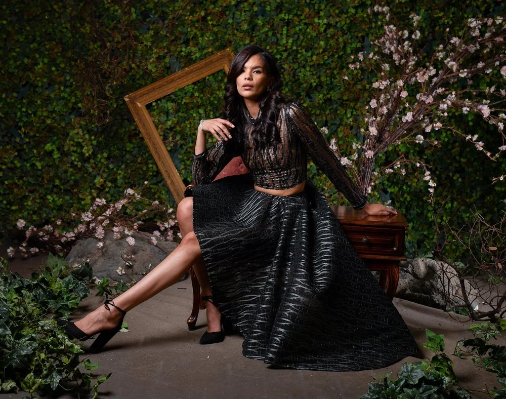 A woman sitting on a wooden table outdoors, surrounded by greenery and pink flowers, with a large wooden frame and rocks in the background. She is wearing a black patterned long-sleeve top and a long black skirt, with black high heels, and has long dark hair.