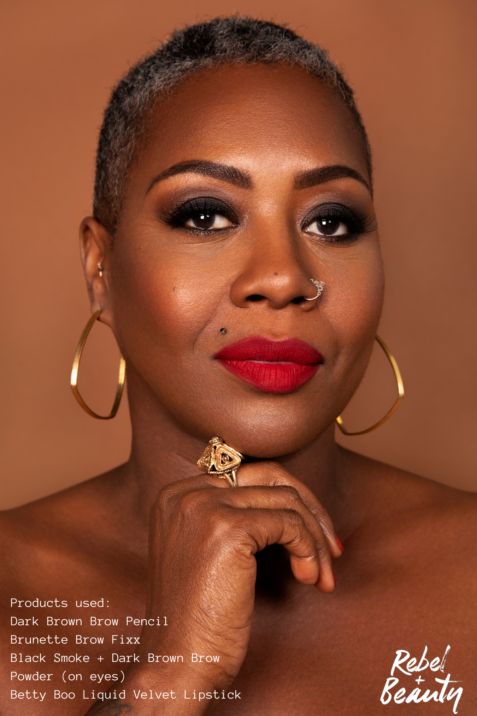 Close-up portrait of a woman with dark brown skin, short gray hair, bold eye makeup, red lipstick, gold hoop earrings, and various facial piercings, holding her hand near her face. Text overlay mentions makeup products used, such as eyebrow pencils, eyeshadow, lipstick, and powder.