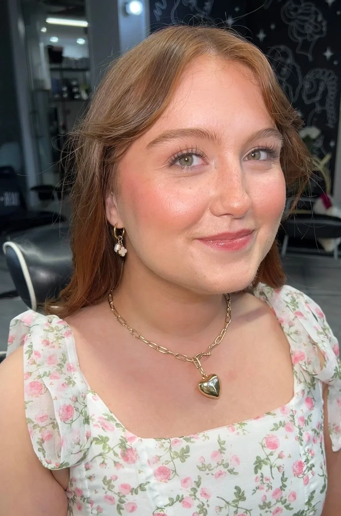Close-up of a woman with reddish-brown hair, wearing a floral dress, gold necklace with heart pendant, and matching earrings, smiling at the camera in a salon or studio.