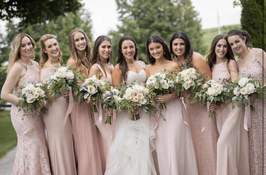 Group of nine women in pastel-colored dresses standing outdoors, holding bouquets of white and pink flowers.
