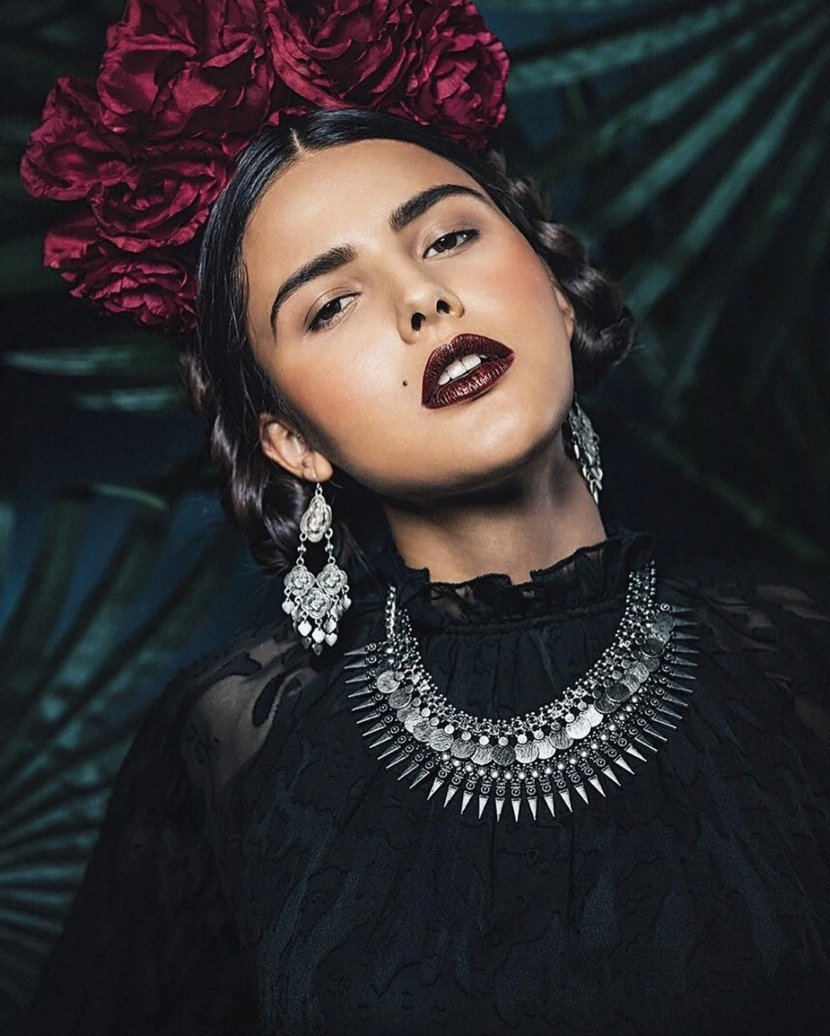 A woman with dark hair styled in waves, wearing dark lipstick, earrings, a layered silver necklace, and a black ruffled top, posed against a tropical foliage background with a large pink flower on her head.