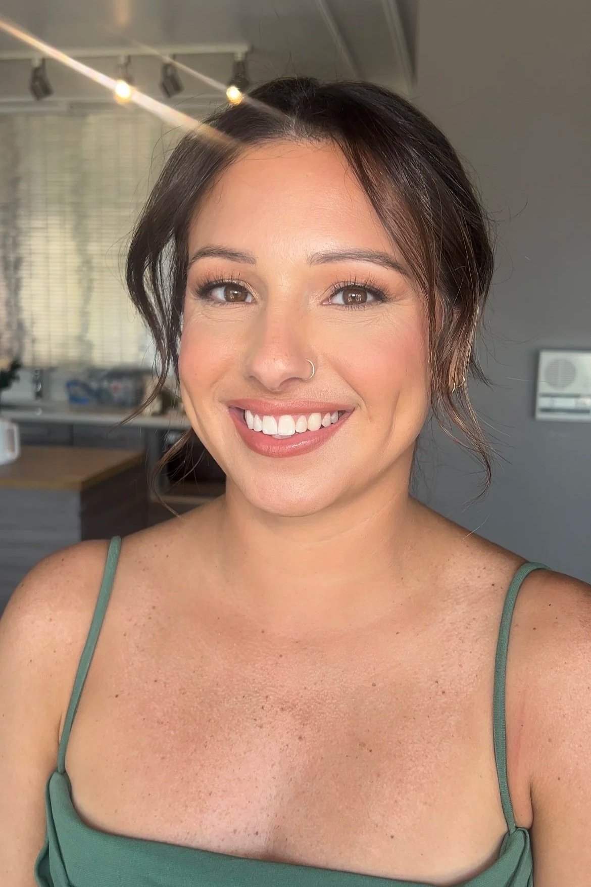 A woman with short brown hair styled in loose waves, wearing a green tank top with thin straps, smiling at the camera inside a room with a gray wall and a window in the background.