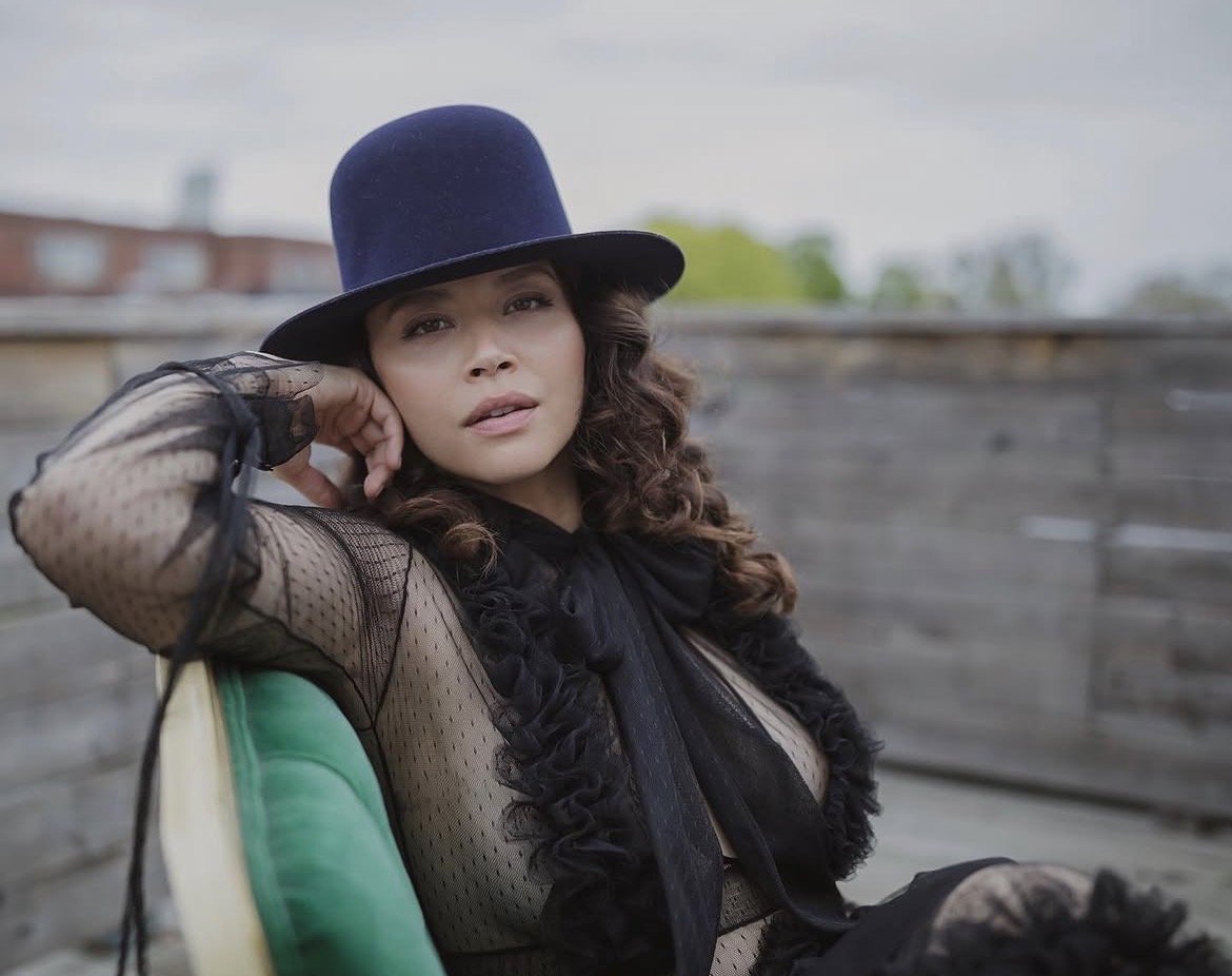 A woman with long curly hair wearing a black top hat and lace mesh top sitting outdoors on a green chair.