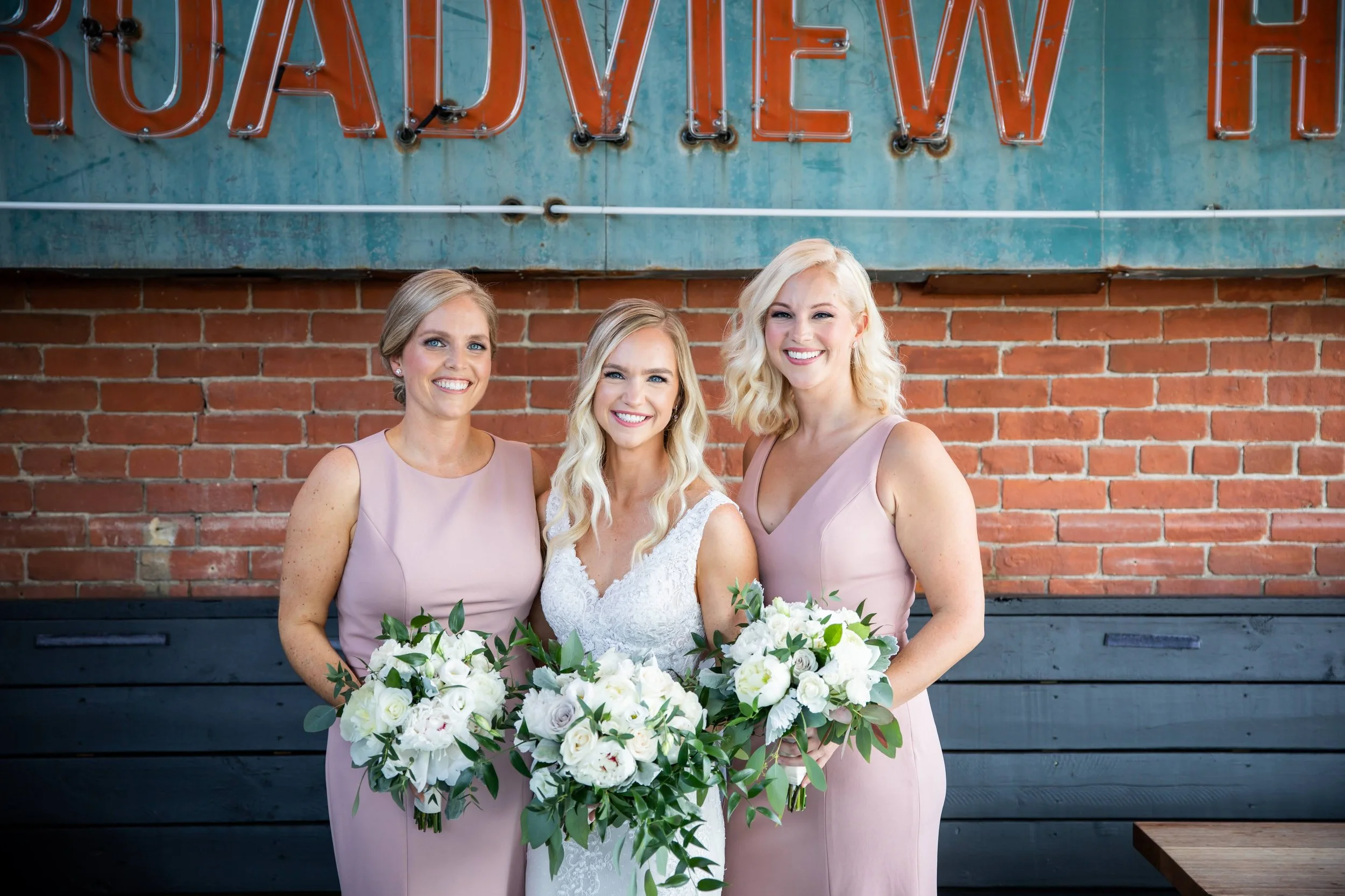 Three women, including a bride in a white wedding dress holding a bouquet, standing in front of a brick wall with a rusted sign that says 'TOURAD VIEW'.