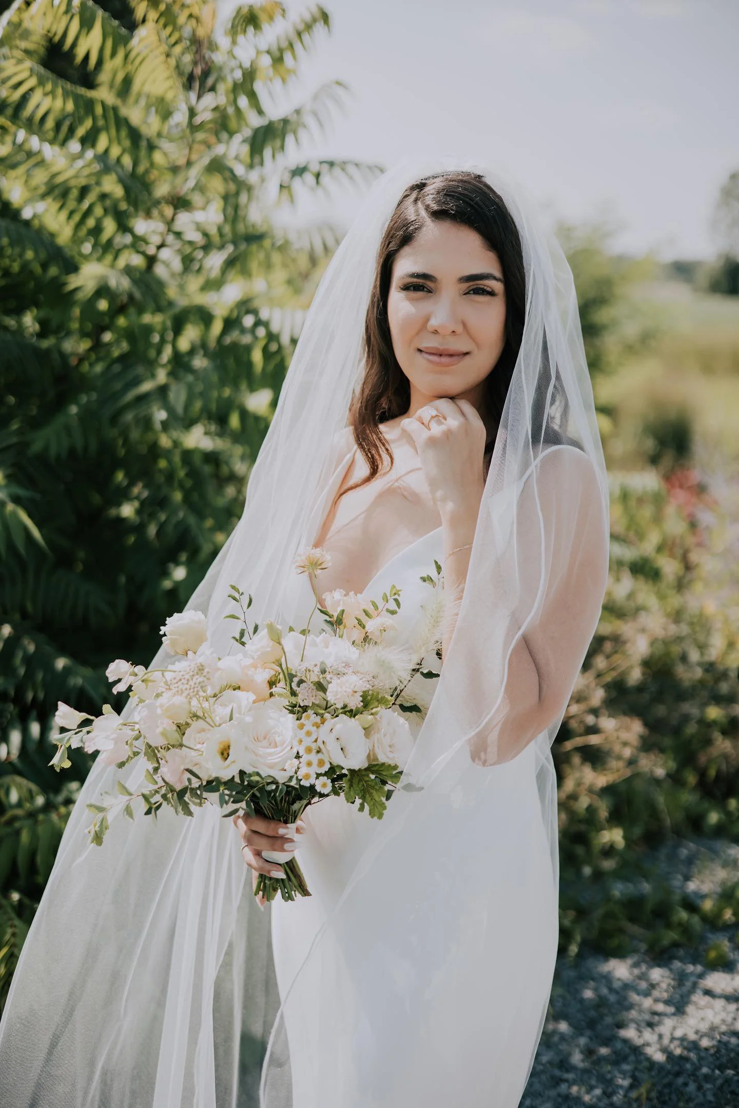 A woman dressed as a bride in a white wedding gown holding a bouquet of white flowers outdoors with greenery and blue sky in the background.