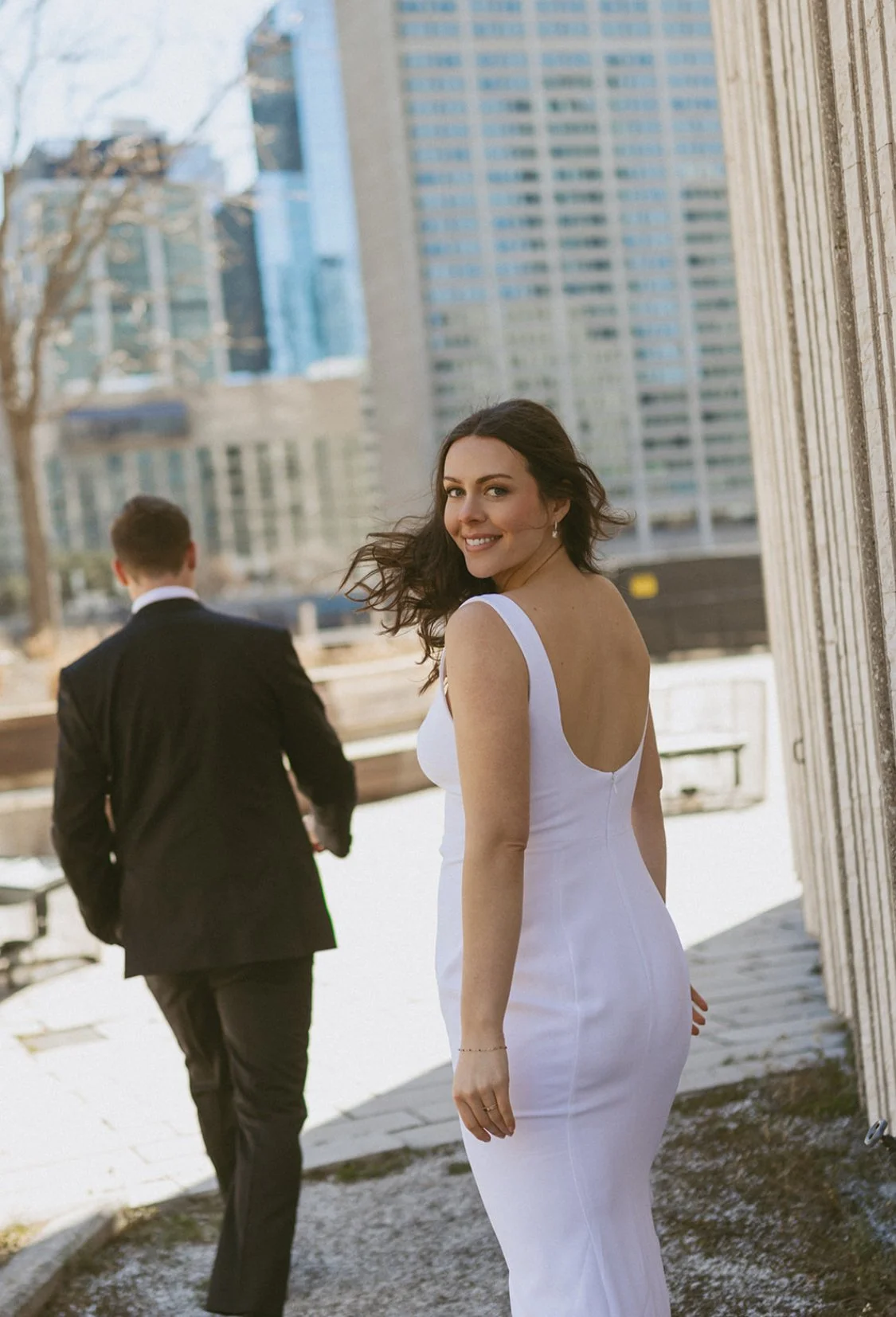 A woman in a white dress and a man in a black suit walking on a city sidewalk, with tall buildings in the background and the woman smiling at the camera.