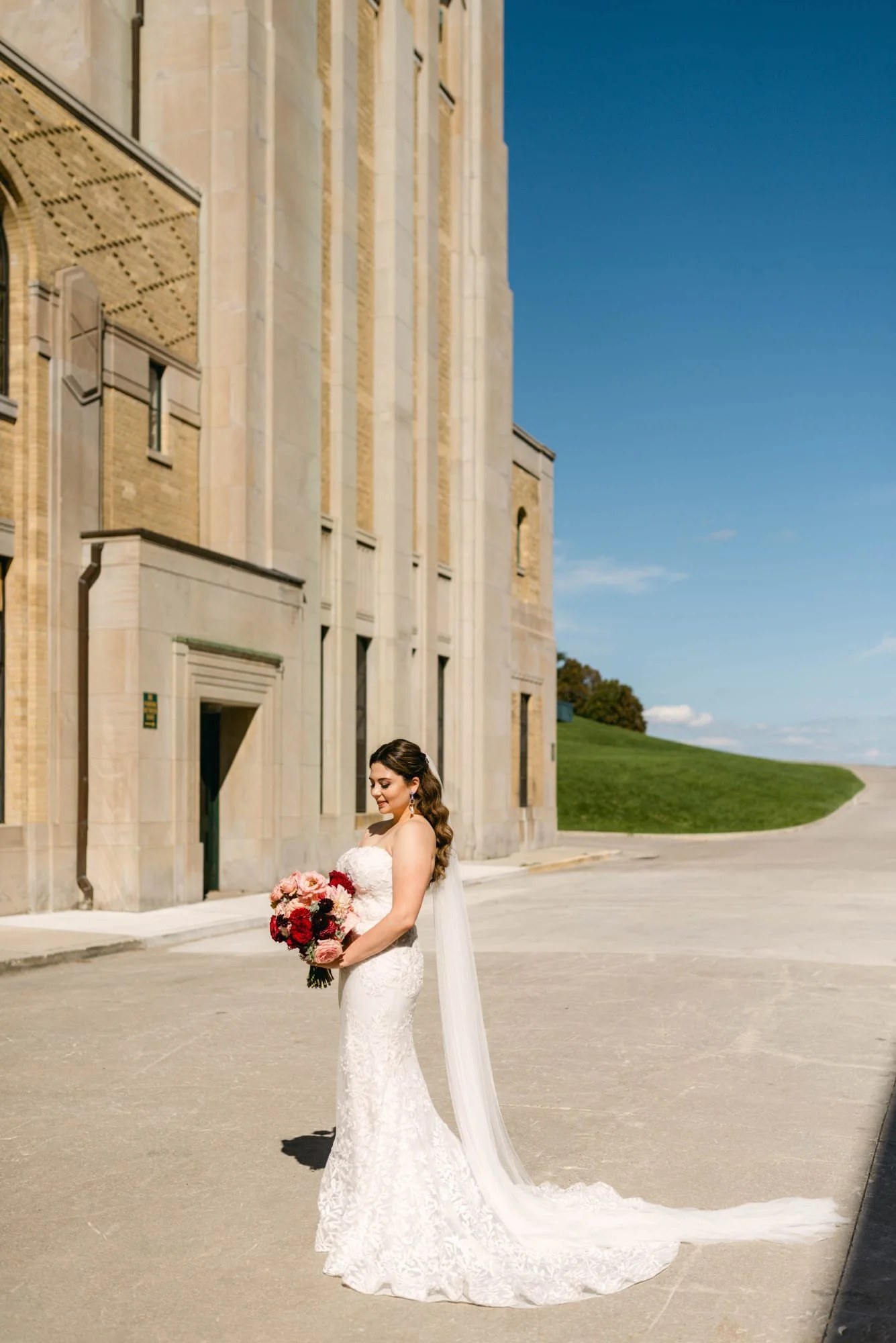 A bride in a white lace wedding dress holding a bouquet of pink, red, and white flowers, standing outdoors in front of an ornate stone building with a blue sky background.