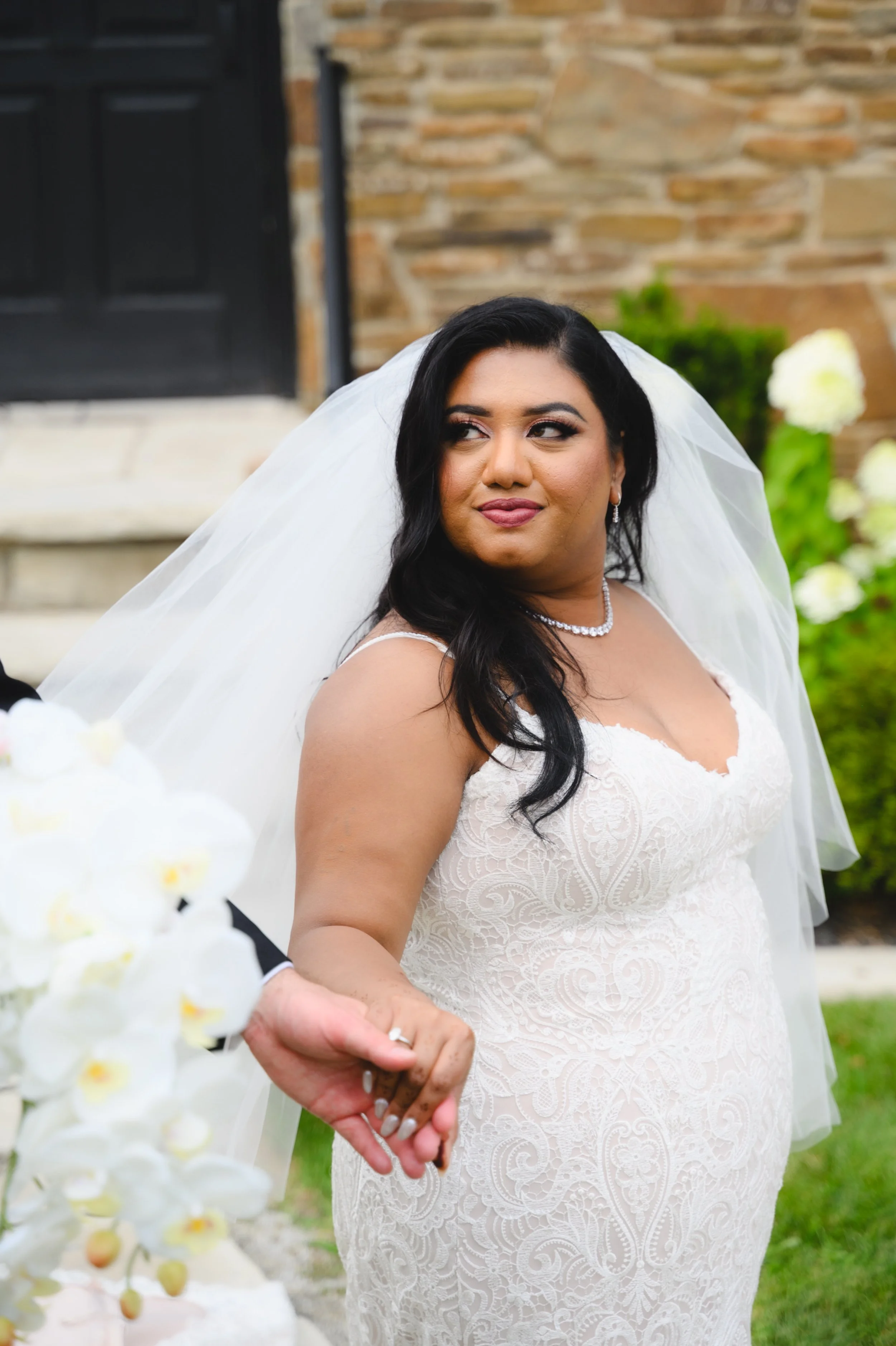 A bride with long black hair wearing a white lace wedding dress, pearl necklace, and drop earrings, holding hands with someone during her wedding ceremony outdoors.