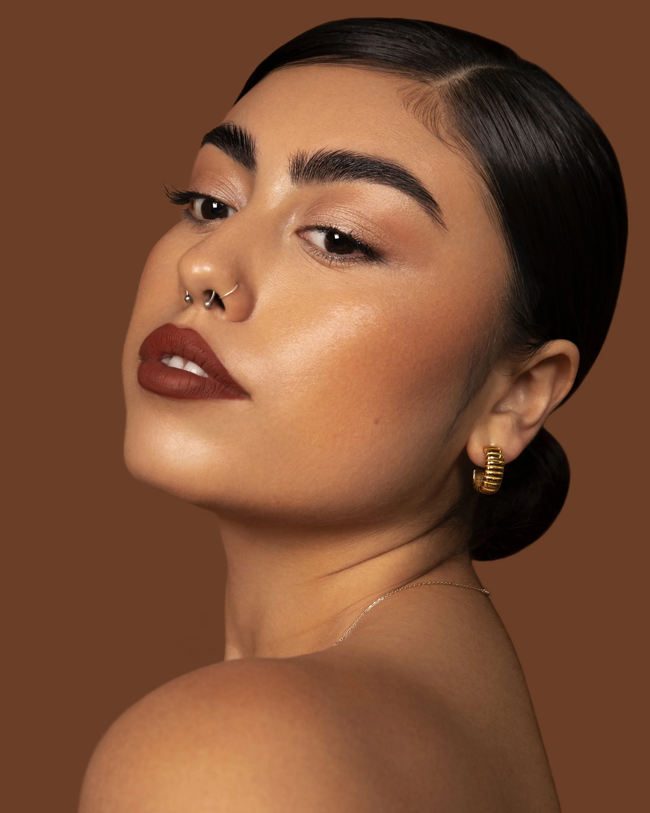 Close-up of a woman's face and shoulder with makeup, dark hair styled sleek and straight, gold hoop earrings, against a plain brown background.