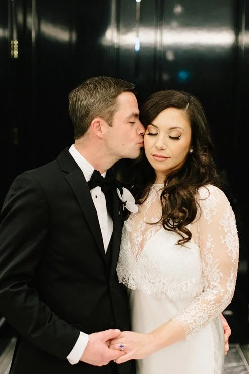 A man in a black tuxedo kissing a woman in a white lace dress on the forehead at a formal event.