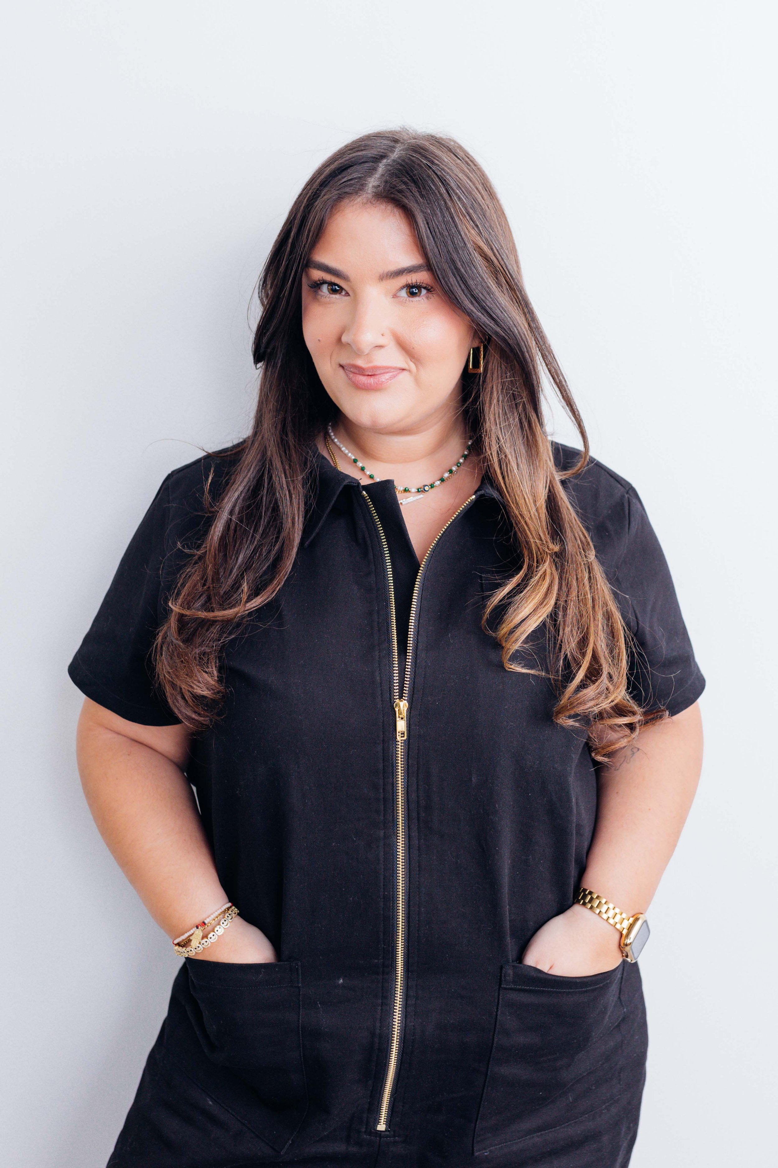 A woman with long brown hair wearing a black zip-up dress, accessorized with jewelry including earrings, necklaces, a bracelet, and a watch, standing against a plain white background.