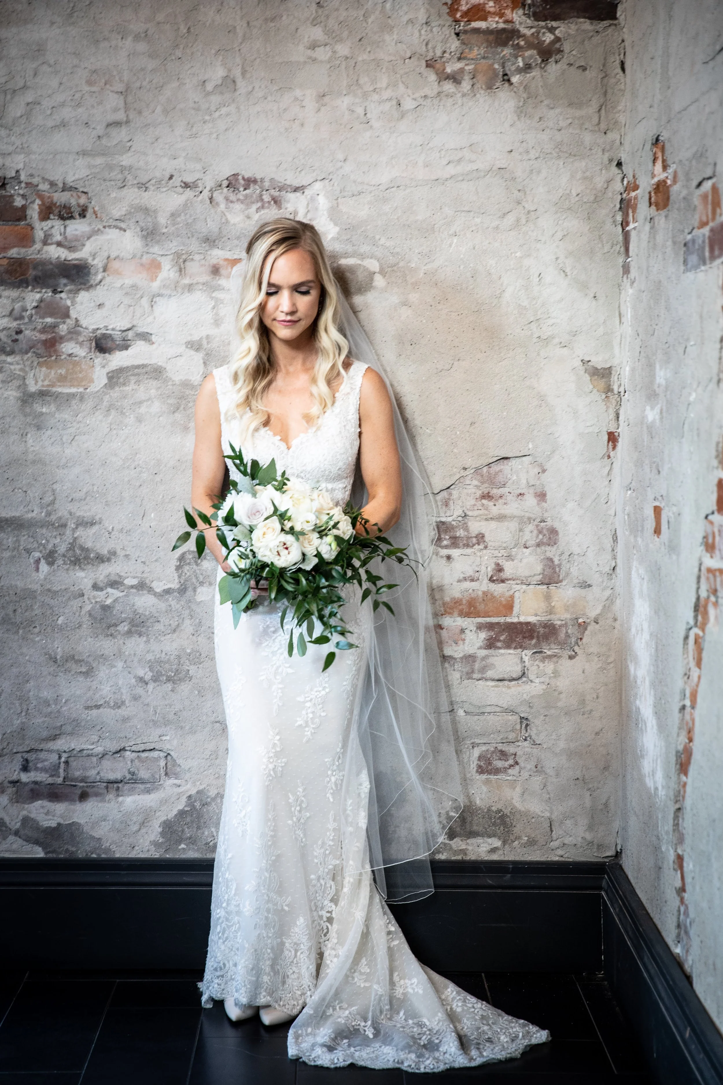 A bride with blonde hair in loose waves, in a white lace wedding gown with a V neckline, holding a bouquet of white roses and greenery, standing against a rustic, exposed brick and plaster wall, with her eyes closed and a serene expression.