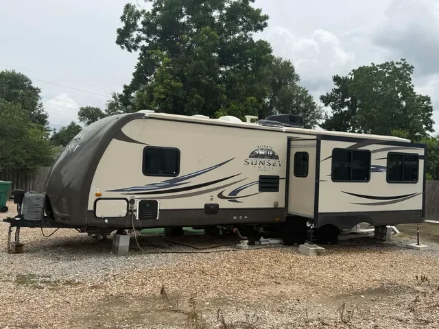 A beige and black Sunset recreational vehicle parked on gravel with trees and cloudy sky in the background.