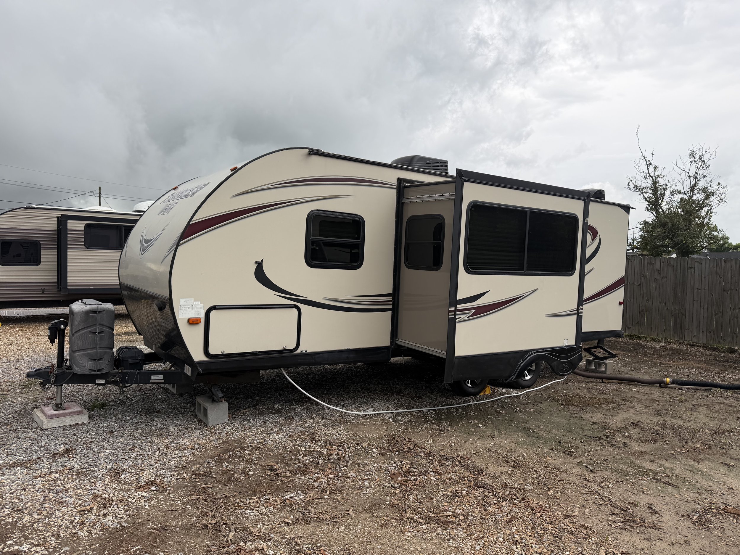 A beige travel trailer with black, red, and gray accents parked on gravel, with overcast skies and trees in the background.