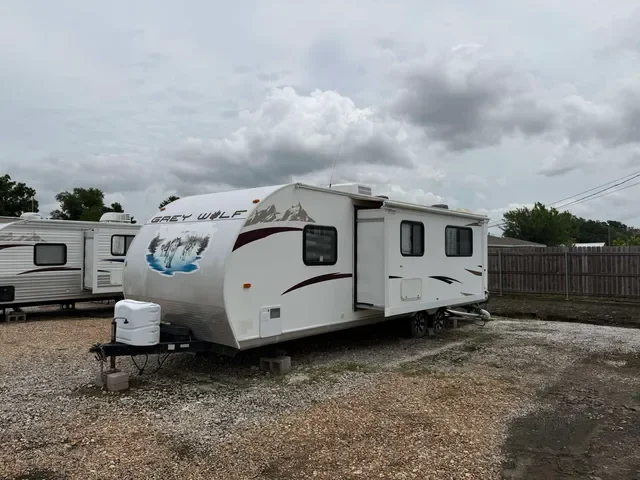 White Grey Wolf travel trailer with an open slide-out section, parked on gravel ground in an outdoor lot, with a wooden fence and cloudy sky in the background.