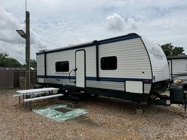A travel trailer parked on a gravel lot with a picnic table and rug outside, under a cloudy sky.