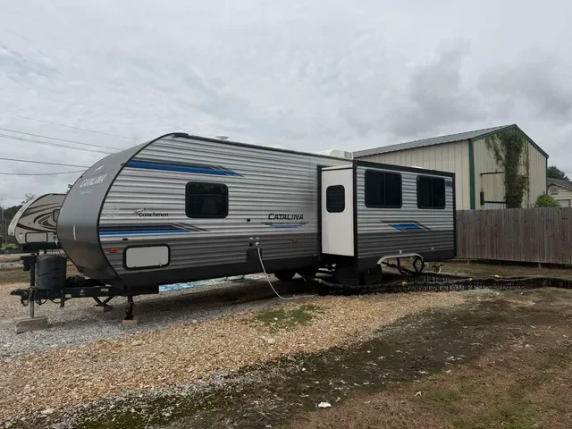 A gray and black Coachmen Catalina travel trailer parked on a gravel lot near a fence and a building under a cloudy sky.