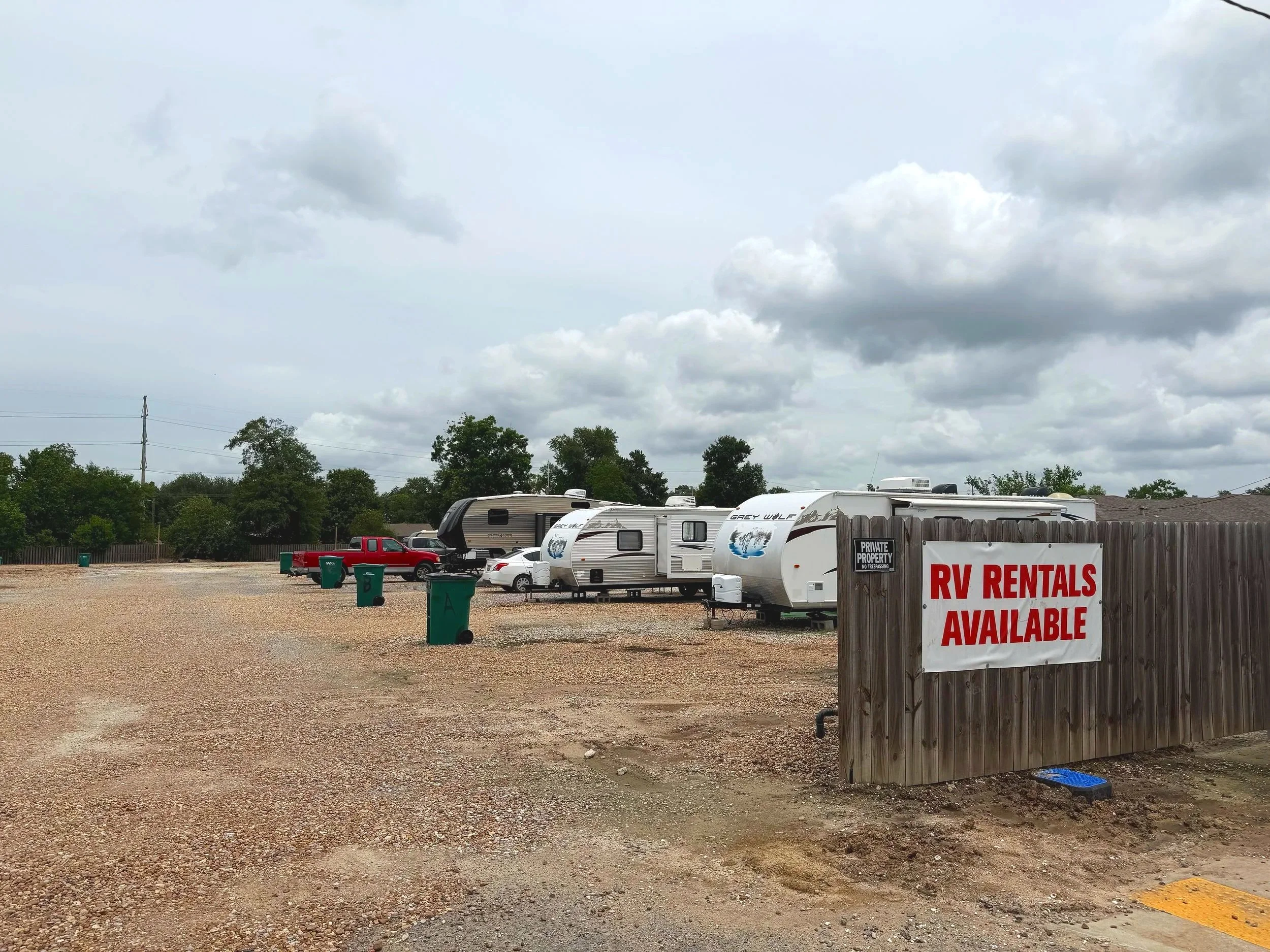 Parking lot with RVs, trucks, and trees under a cloudy sky, with a sign reading 'RV Rentals Available'.