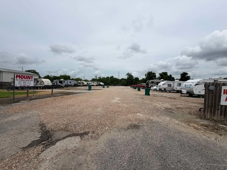 A gravel parking lot with travel trailers and RVs parked along the sides, with a sign that reads 'MOUNT RV & Mobile Home Park' and clouds in the sky.