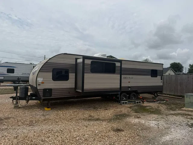A beige and black travel trailer parked on a gravel lot with a wooden fence and other trailers in the background.