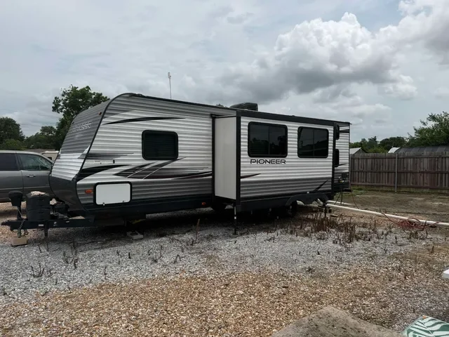 Travel trailer labeled 'Pioneer' parked on gravel with a wooden fence and cloudy sky in the background.