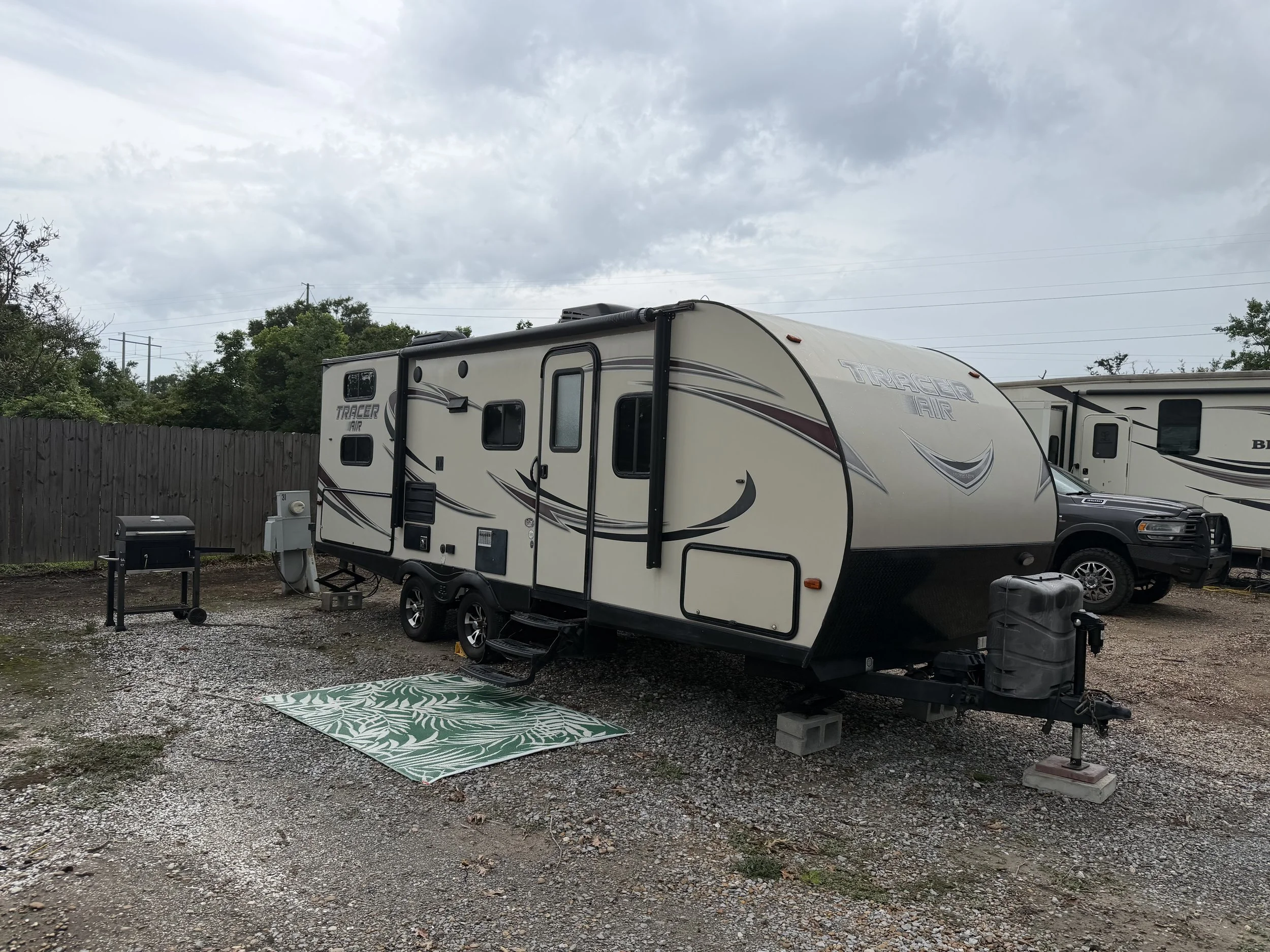 A recreational vehicle (RV) parked on a gravel lot behind a wooden fence, with a black pickup truck parked beside it under a cloudy sky.