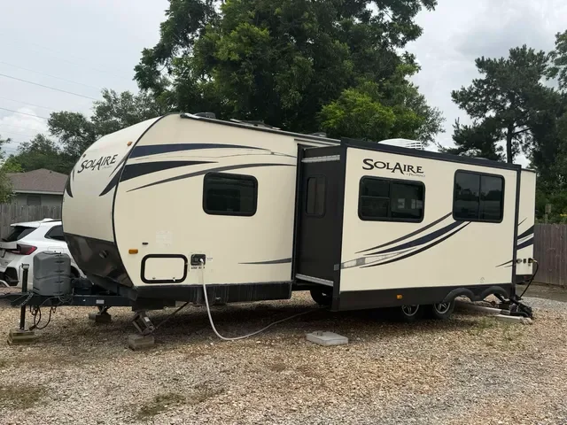 A white SolAire travel trailer with black and gray accents parked on gravel, with trees and a fence in the background.