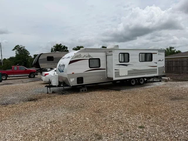 Travel trailer parked on gravel with a red pickup truck and trees in the background under cloudy sky.