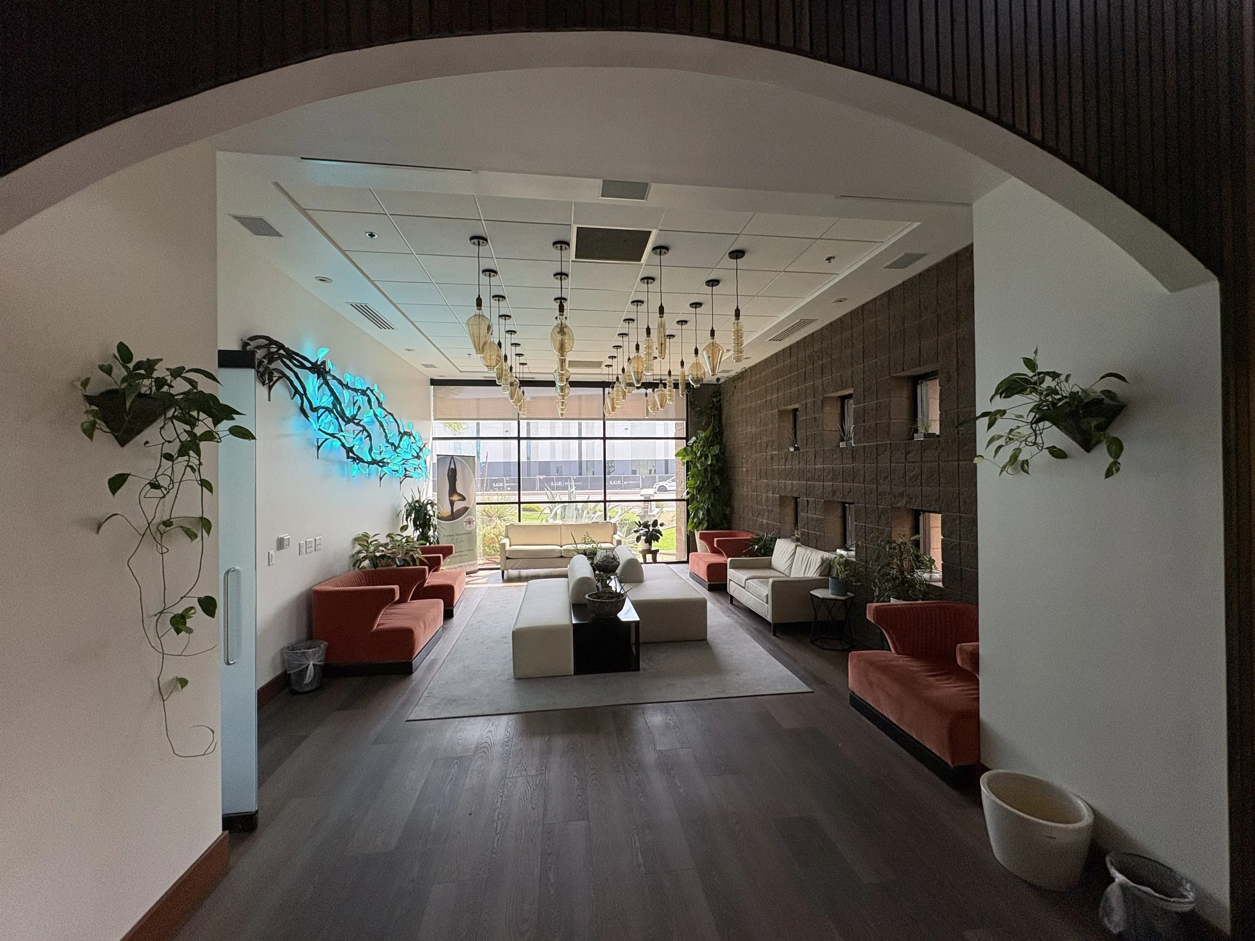 A spacious lounge area with large windows, seating including sofas and armchairs, plants, hanging light fixtures, and a decorative wall with a blue glow, viewed through an arched doorway.
