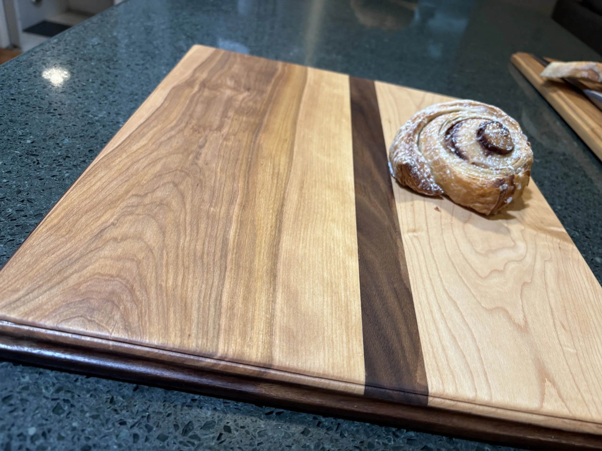 Close-up of a wooden cutting board with a cinnamon roll on it, placed on a green countertop.