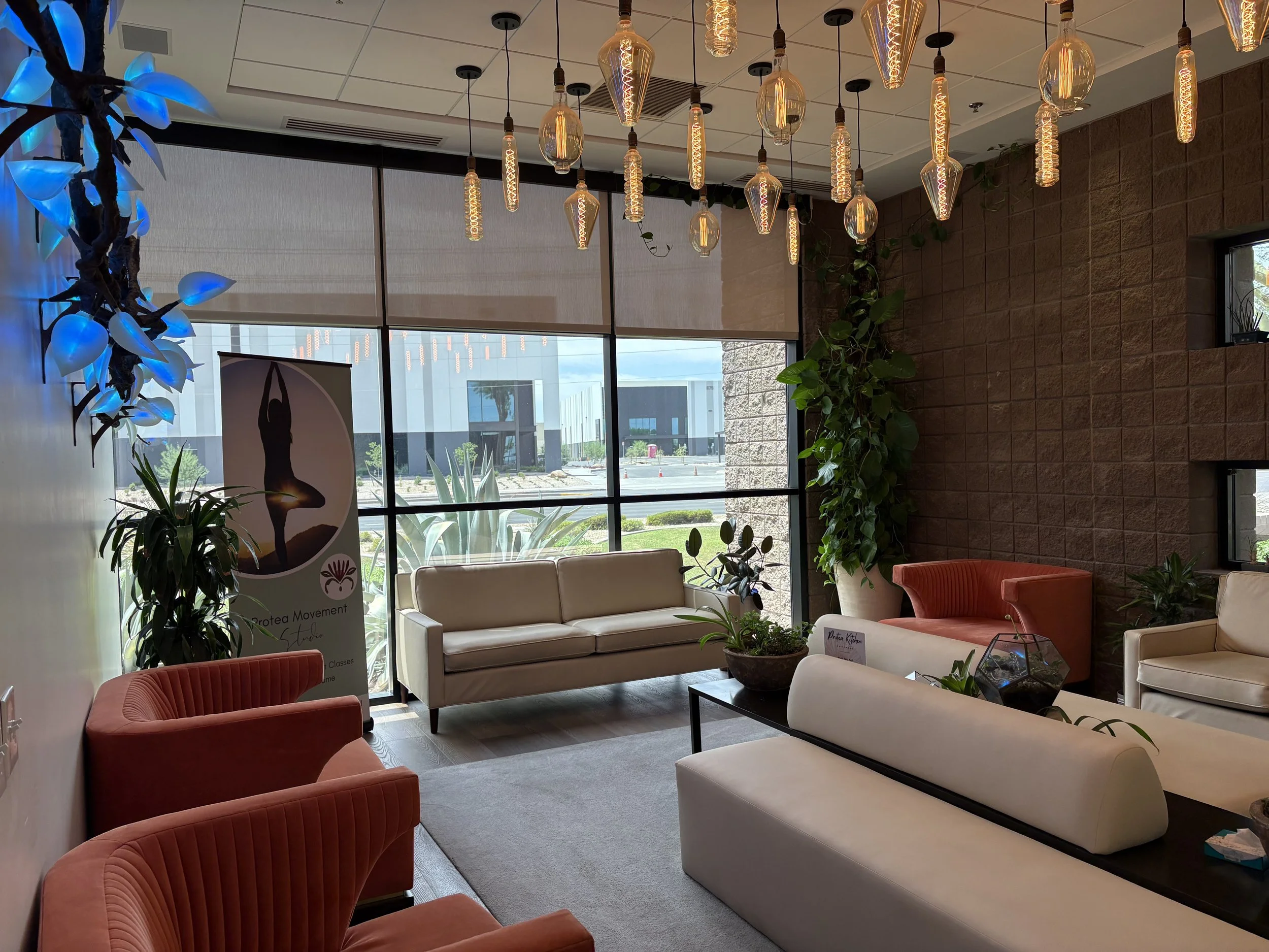 Interior of a modern lounge or waiting area with beige and red seating, large windows, hanging pendant lights, and indoor plants.