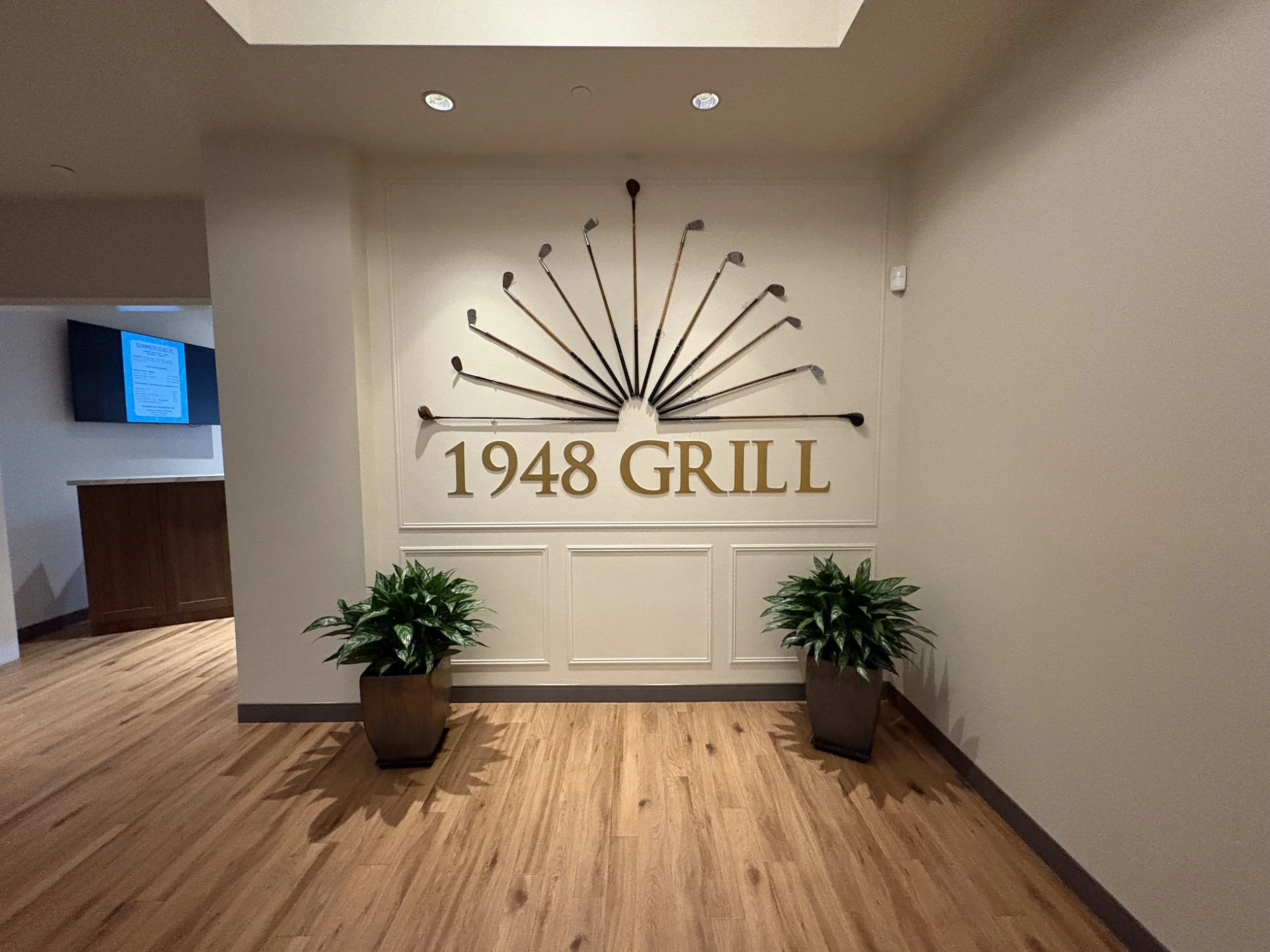 Wall sign with gold text reading '1948 GRILL' and decorative golf clubs above. Two potted plants are on either side of the sign, with wooden flooring underneath.
