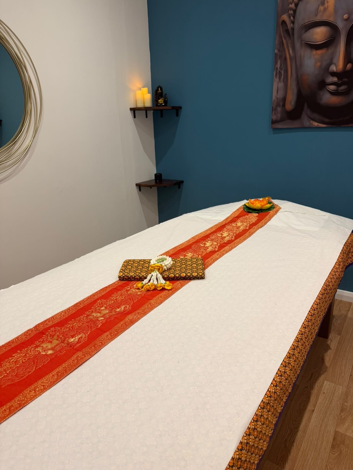 Spa room with a massage table covered in a white sheet, decorated with an orange and gold ribbon, flowers, and small blankets, with candles and a Buddha painting in the background.