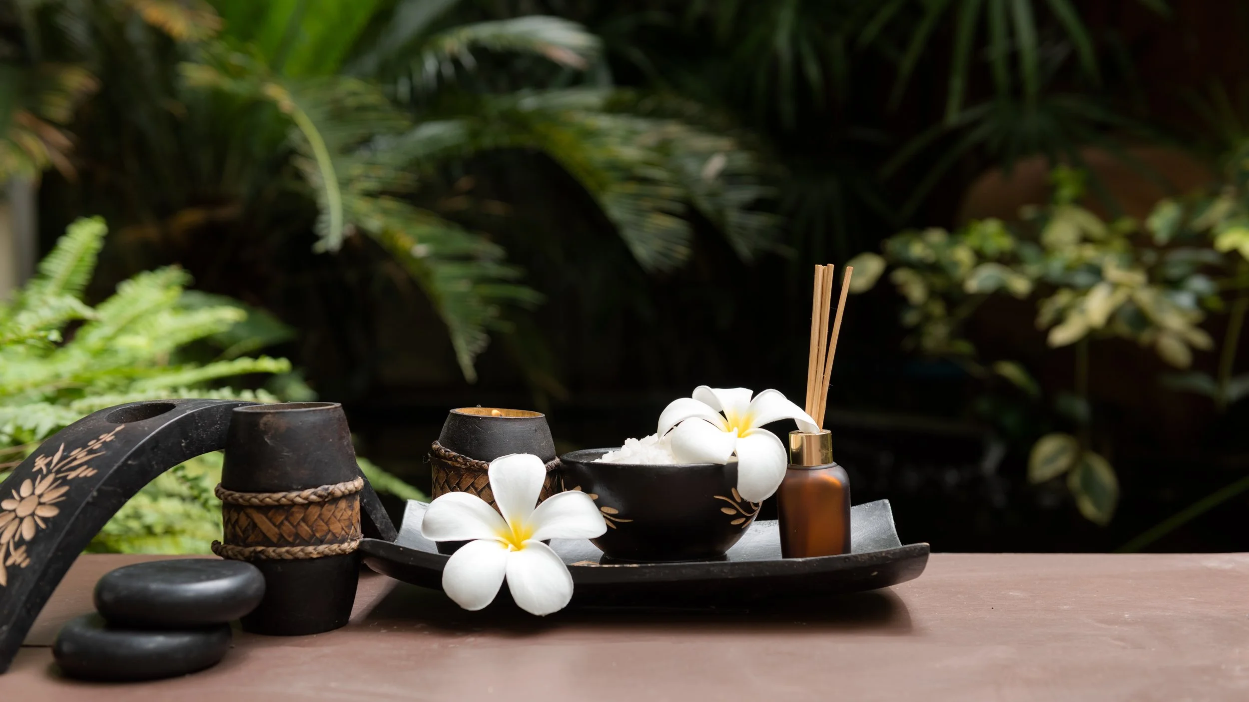A spa setting with black ceramic bowls, white flowers, bamboo brush, and candles on a black tray in a lush green outdoor environment.