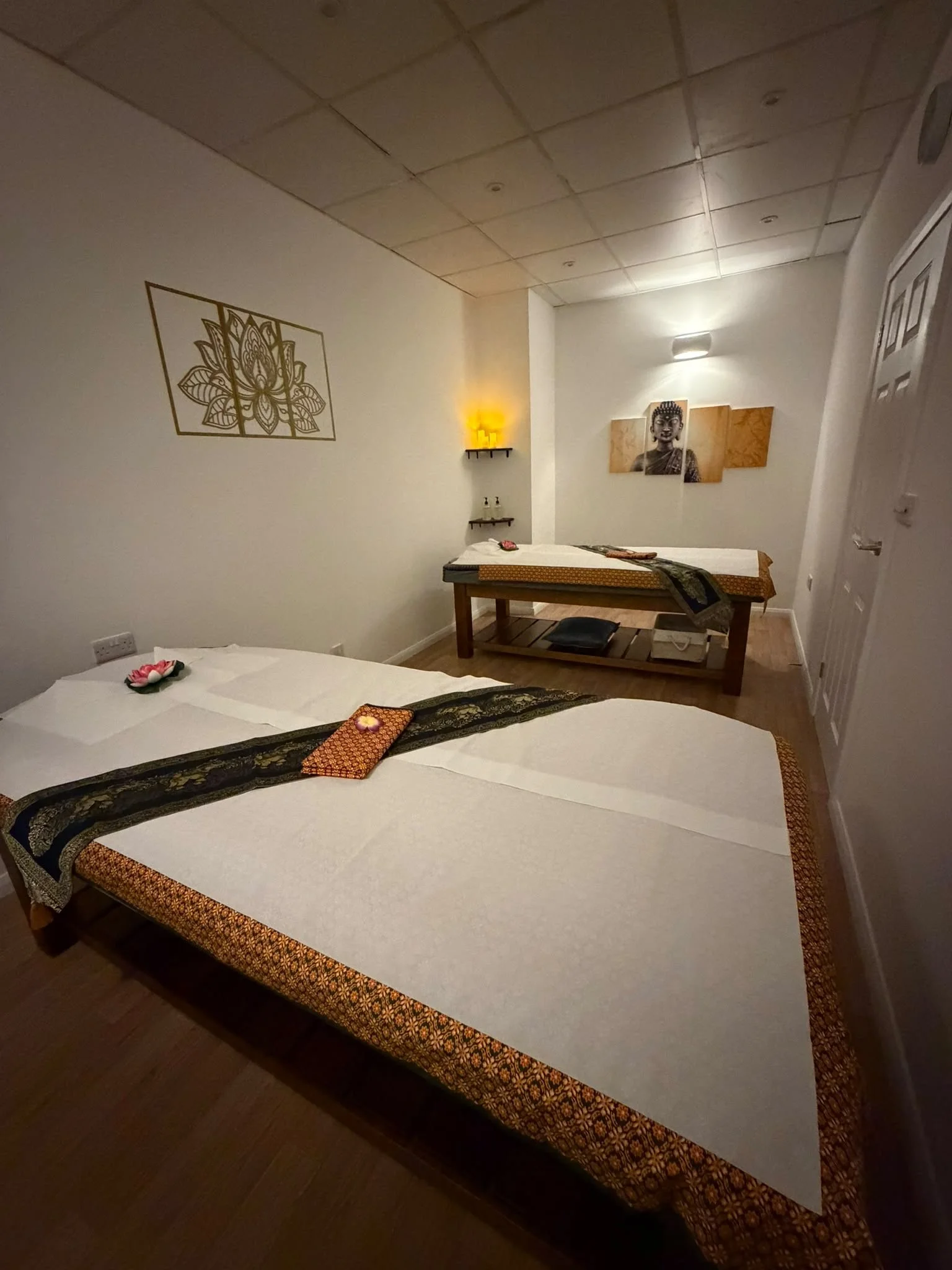 A wellness or massage room with two massage beds decorated with flowers and towels, framed artwork of a deity on the wall, and dim lighting with warm accent lights.