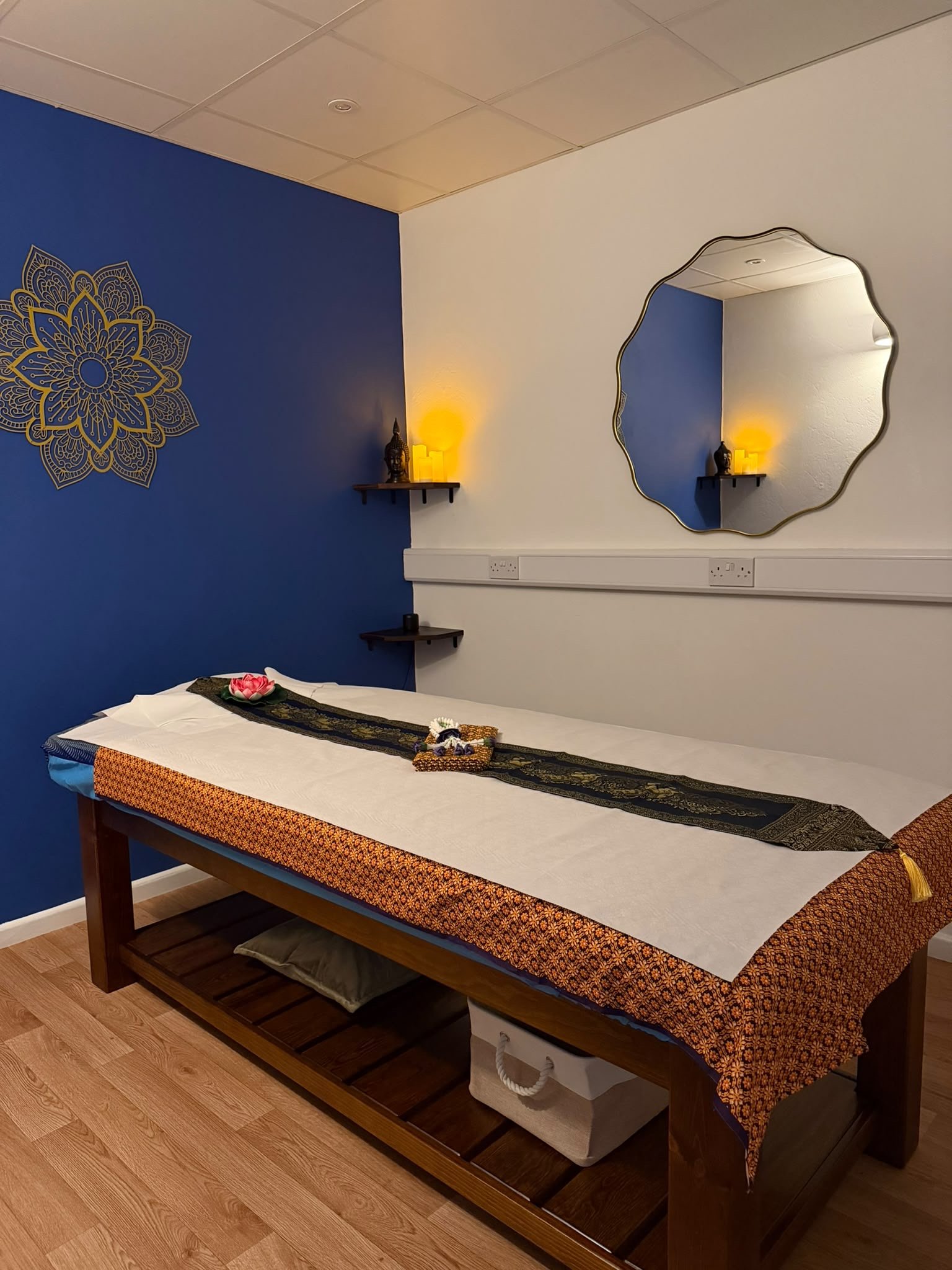 A massage or therapy room featuring a massage table draped with a patterned cloth, a lotus flower and small decor items on top, a mirror on the wall reflecting the room, a floating shelf with candles and a Buddha statue, and a dark wood and blue accent wall with a decorative pattern.