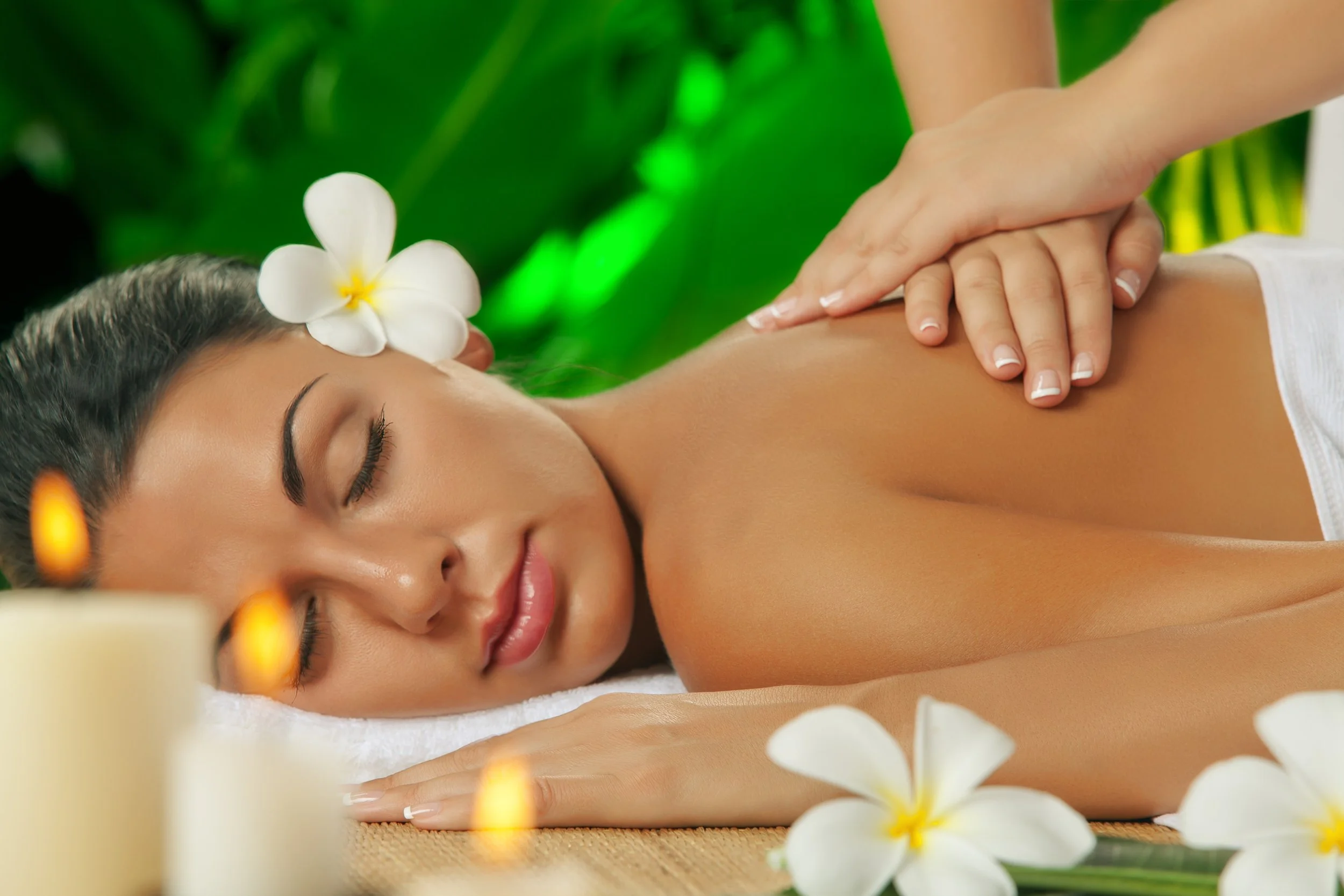 A woman receives a massage at a spa, with white plumeria flowers and candles in the foreground.
