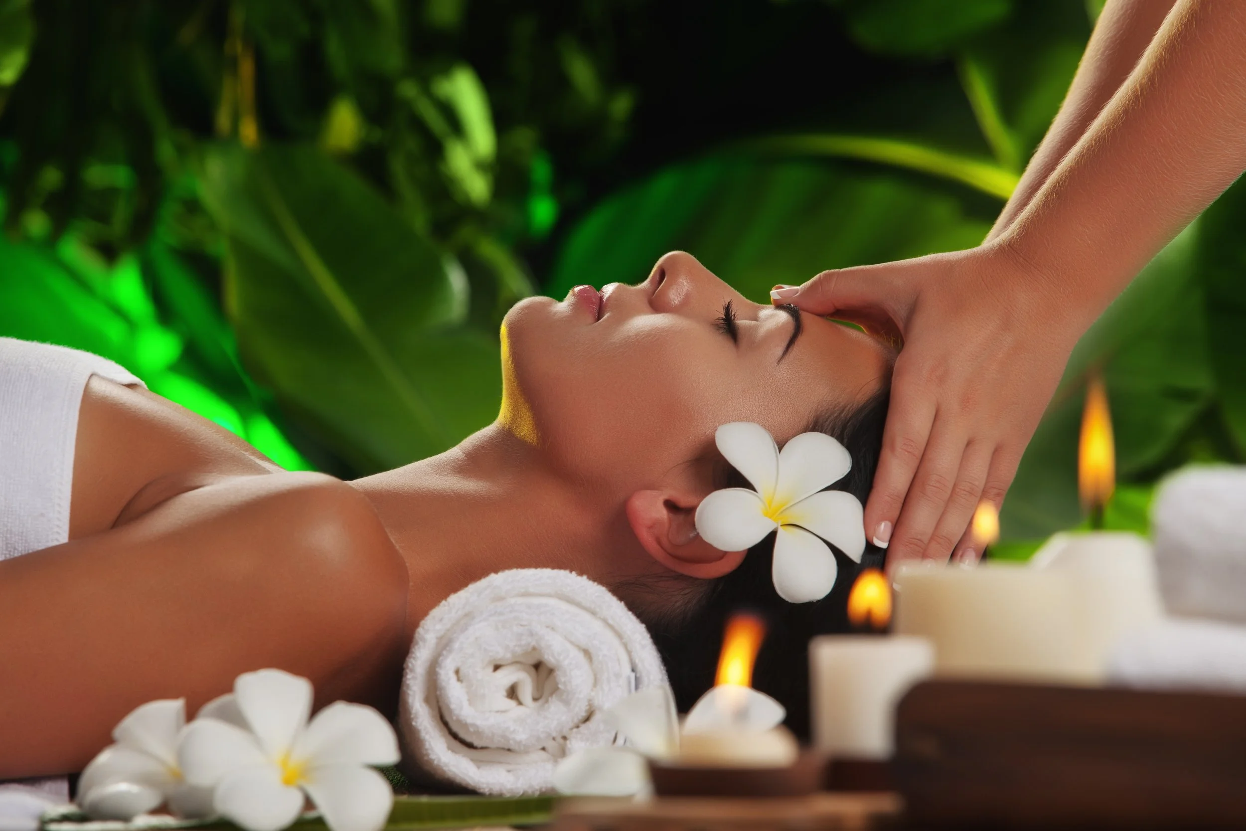 A woman receiving a massage on her neck in a spa, surrounded by white flowers, rolled towels, and lit candles, with lush green plants in the background.