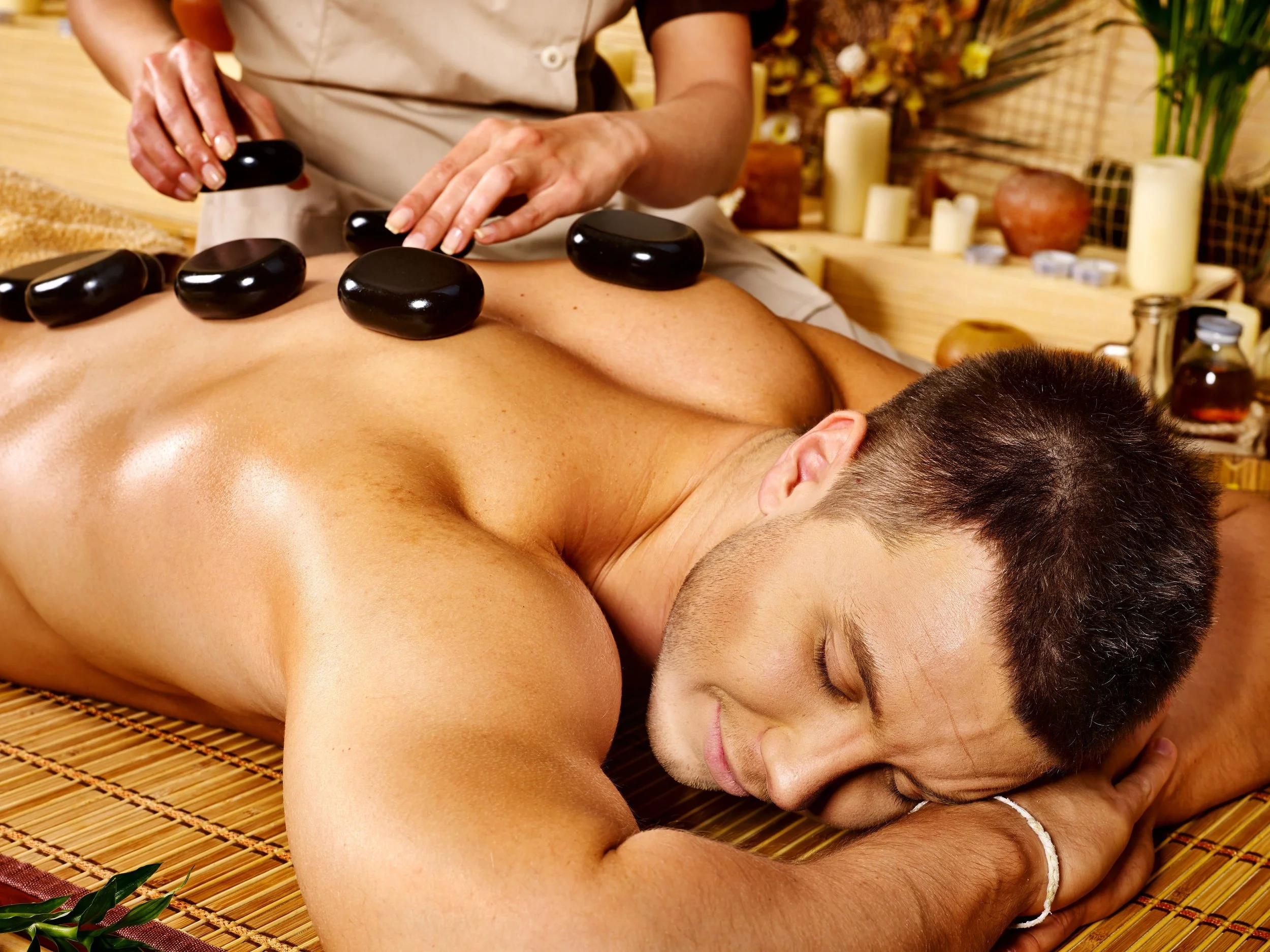 A person receiving hot stone massage therapy on their back, lying face down on a mat with black stones placed along the spine, in a serene massage room with candles and decorative plants.